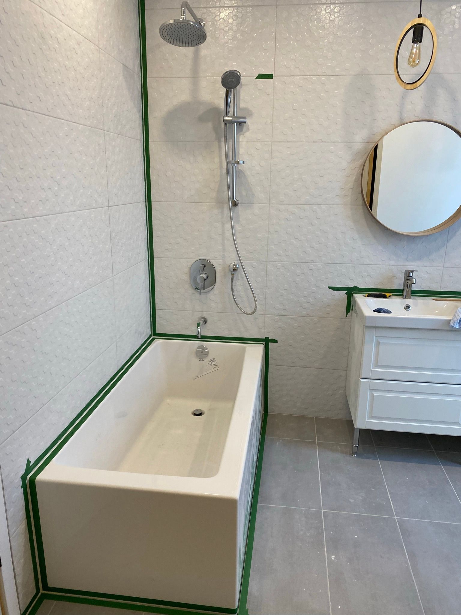 Rénovation de salle de bain avec carrelage blanc, baignoire, robinetterie de douche, meuble-lavabo et miroir rond. Ruban adhésif vert délimite les murs.