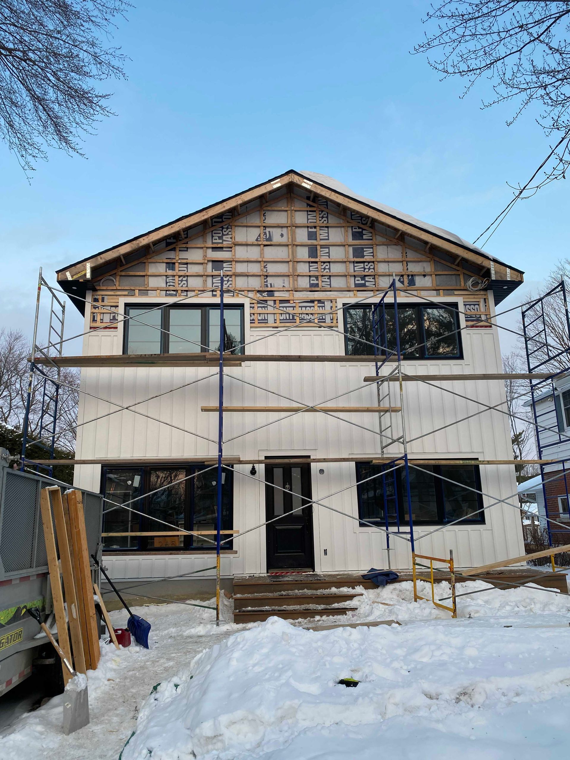 Maison à deux étages en construction. Échafaudages, bardage blanc, fenêtres noires, neige au sol, ciel bleu.
