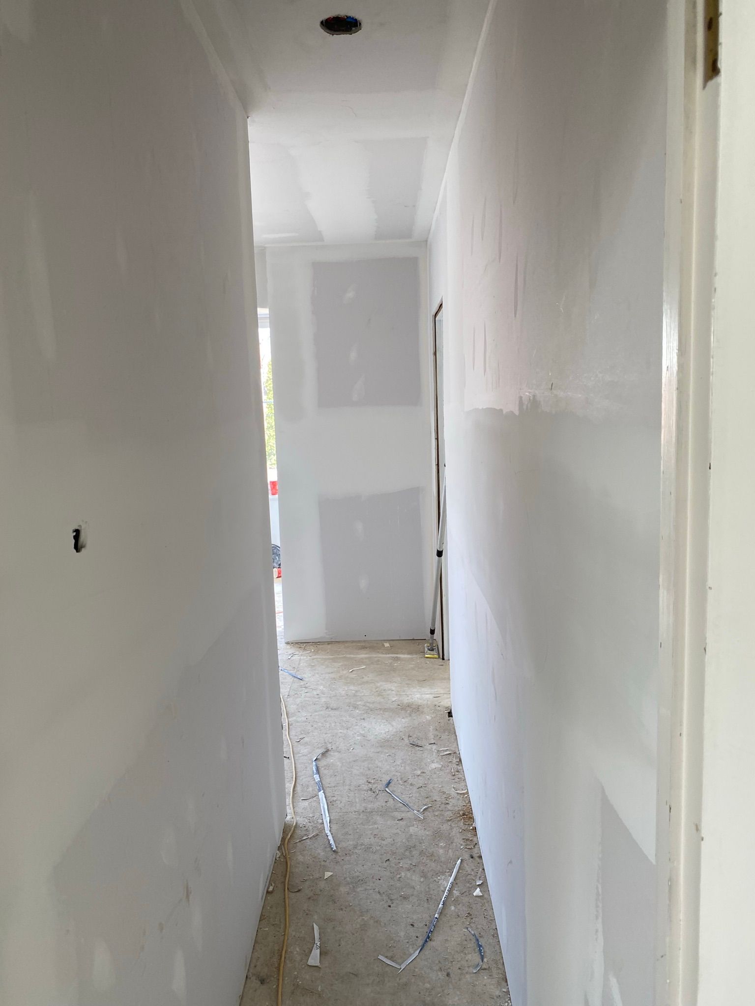 Couloir étroit en travaux ; cloisons sèches, sol non fini, porte ouverte au fond.