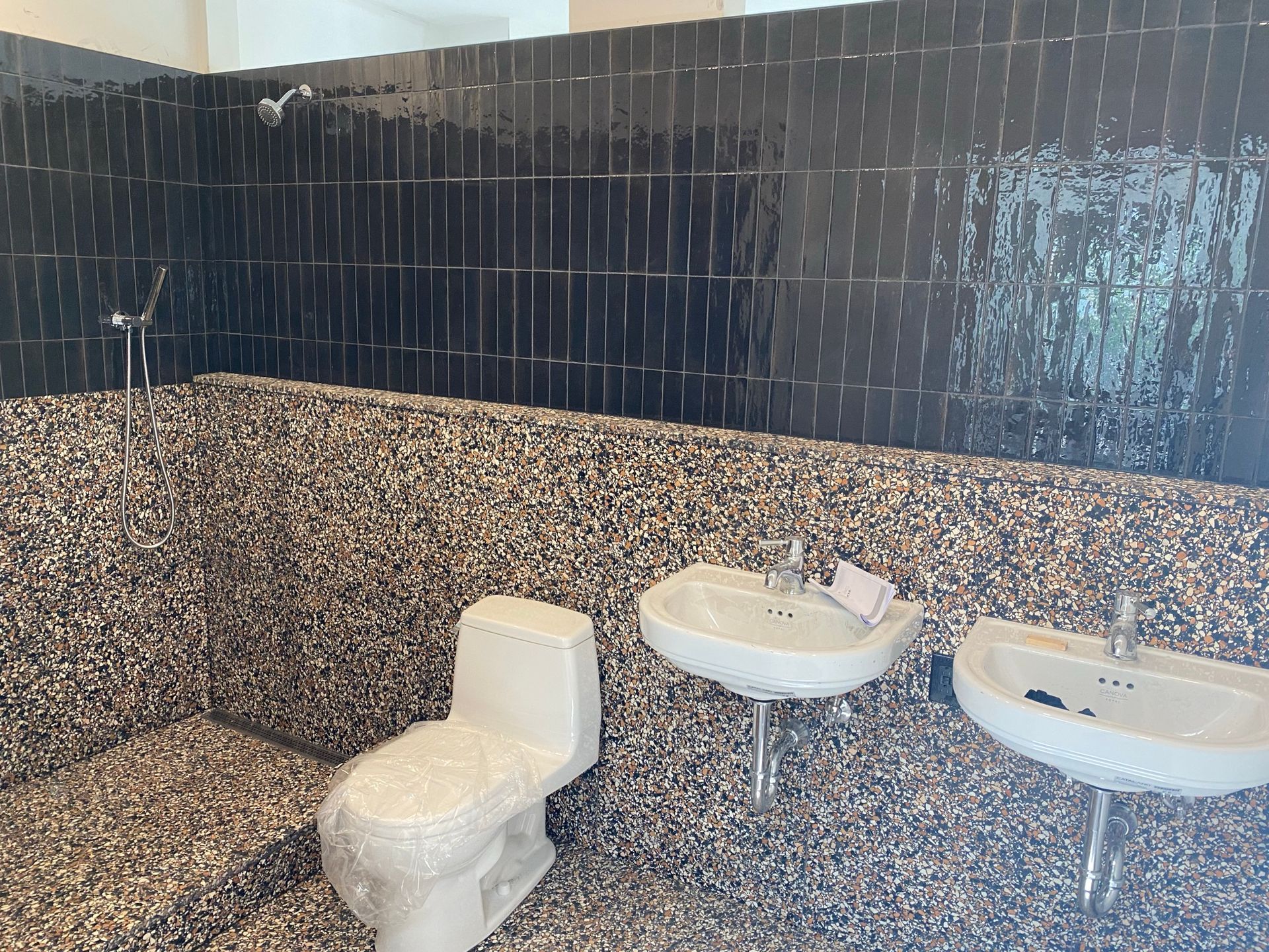Salle de bains avec douche carrelée de noir et murs recouverts de galets. Deux lavabos et des toilettes sont visibles.