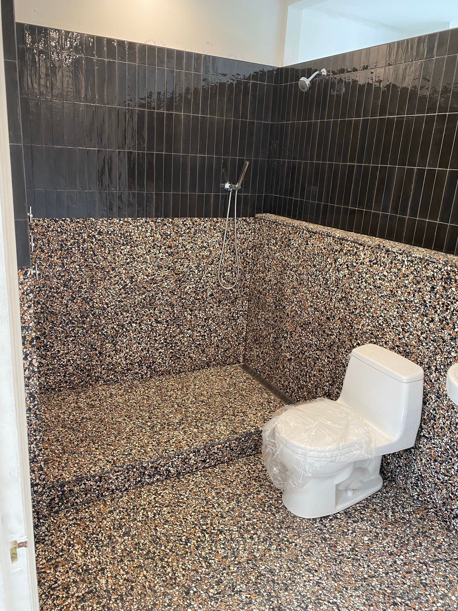 Salle de bains avec douche carrelée de noir et carrelage mural et au sol moucheté, toilettes.