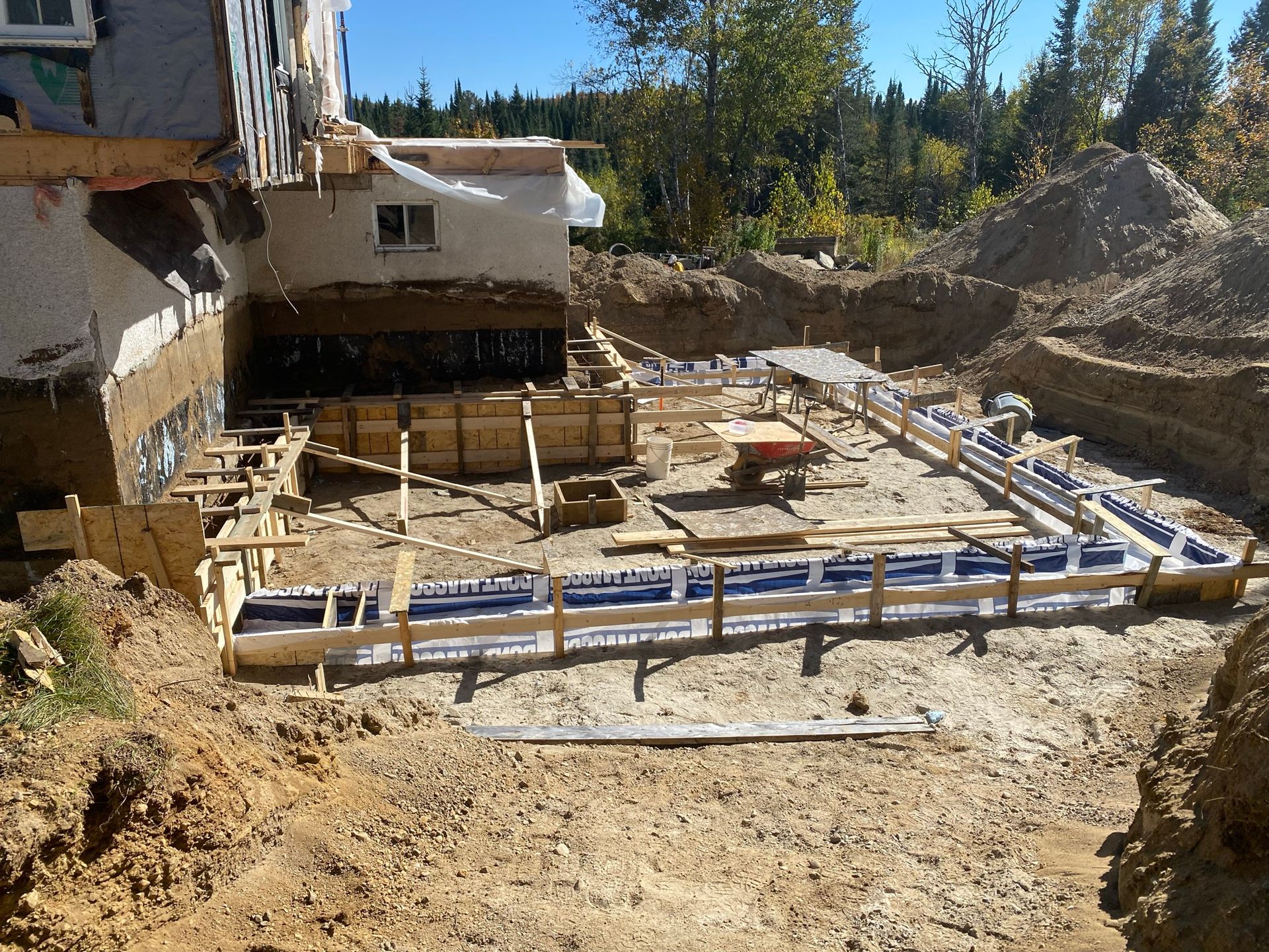 Chantier : Coffrages en bois pour fondations, fixés à une structure existante, installés dans la terre excavée.