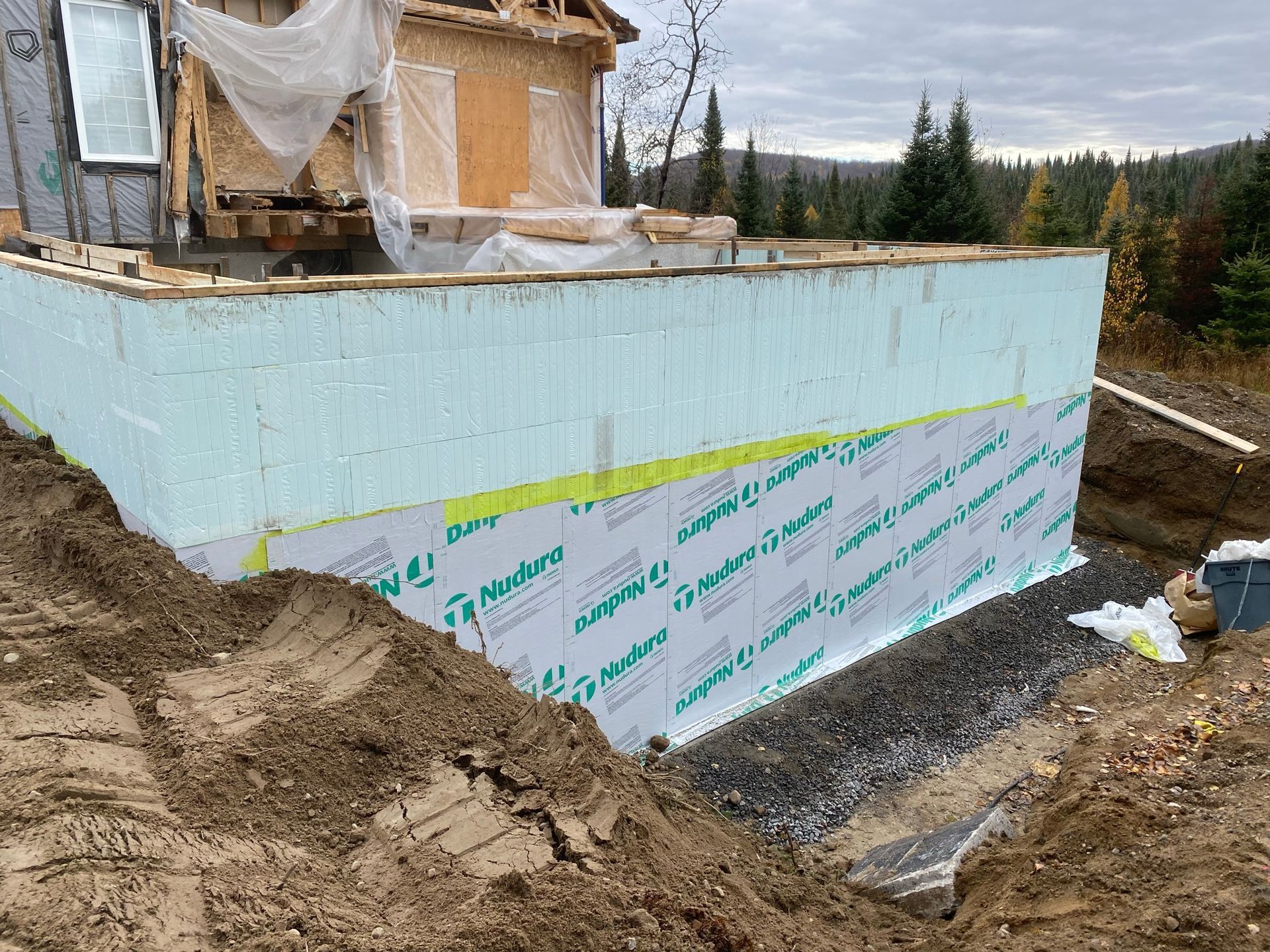 Chantier : Mur de fondation avec isolation bleue et étanchéité verte, terre et gravier visibles.