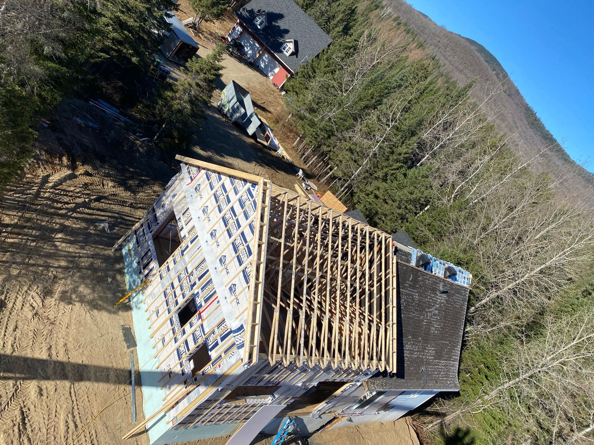 Vue aérienne d'une maison en construction ; charpente en bois apparente, entourée d'arbres et d'une montagne.