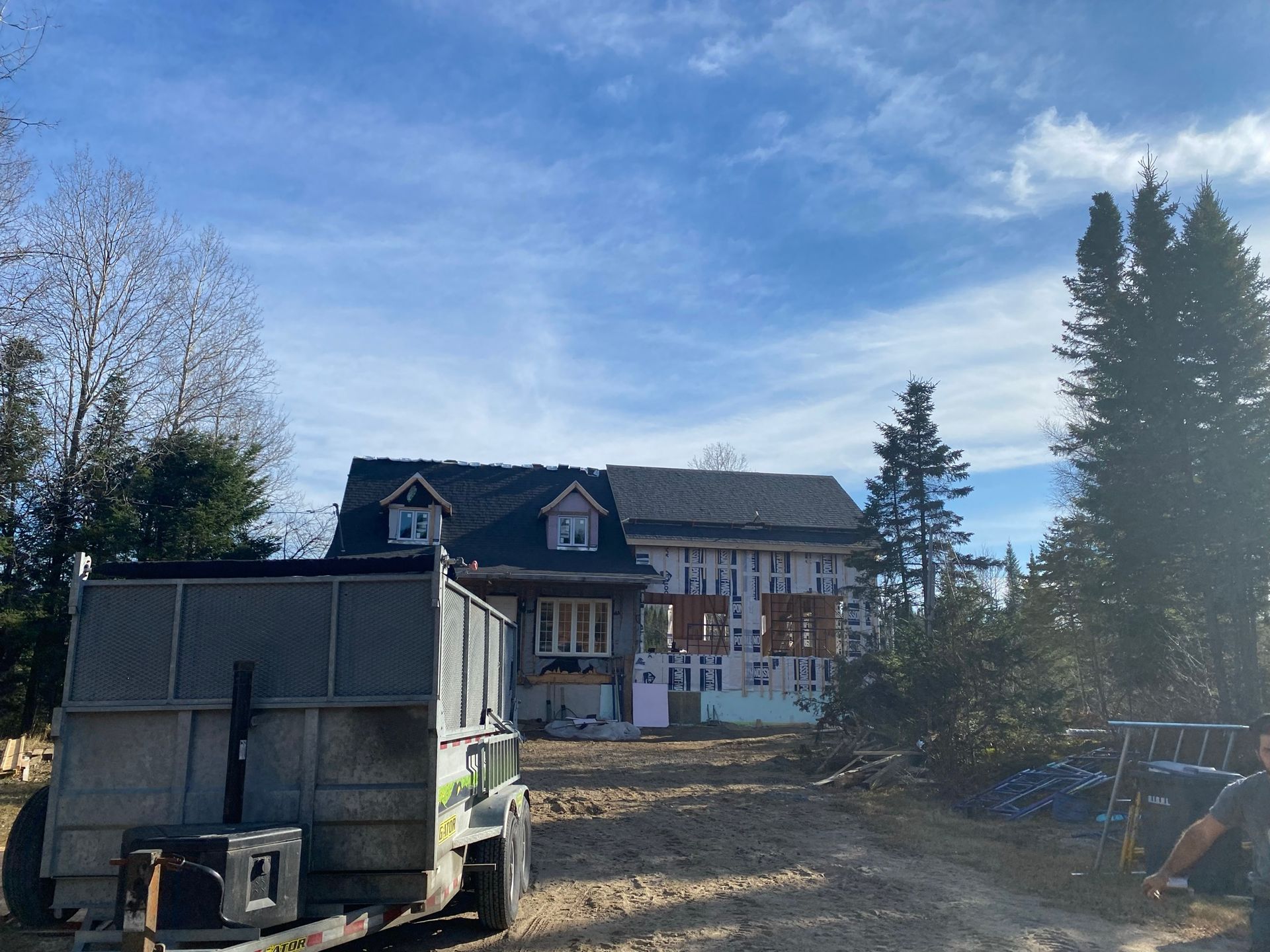 Chantier : maison à la structure partiellement construite, toit noir, ciel bleu et une caravane au premier plan.