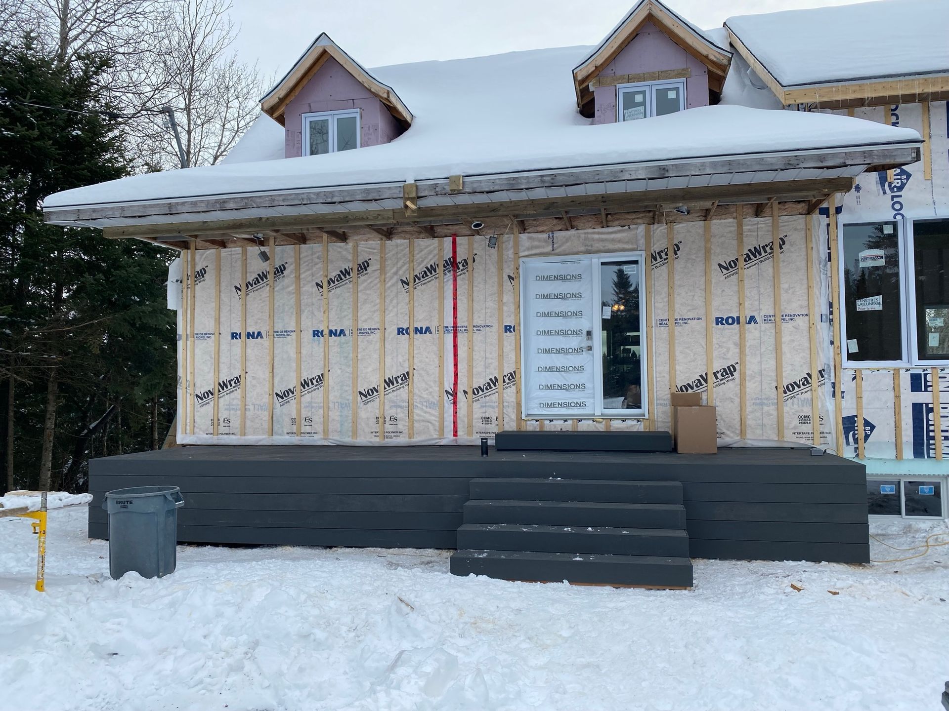 Extérieur d'une maison en construction en hiver, avec un toit enneigé, des murs inachevés et une plateforme grise.