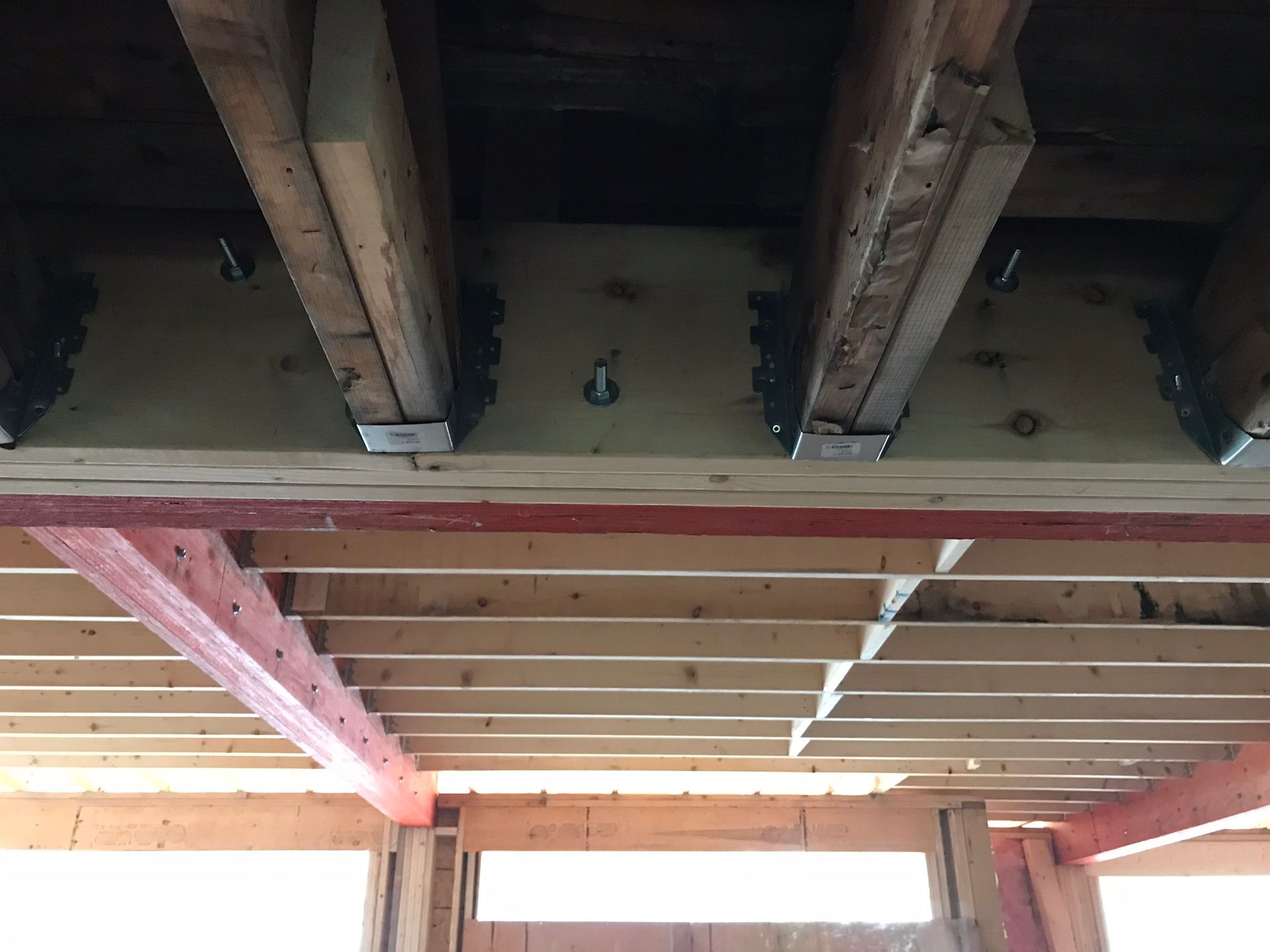 Vue de dessous de la structure en bois de la terrasse : poutres, supports et consoles. Peinture rouge visible.