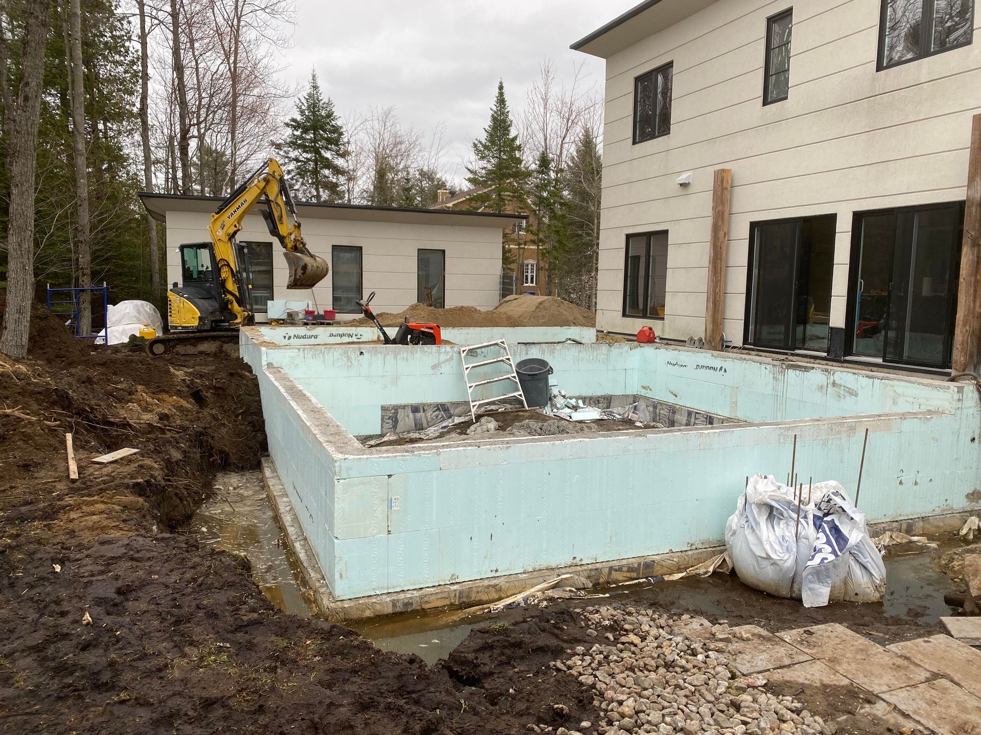 Chantier avec piscine et bâtiment inachevés. Pelleteuse, débris et terre. Ciel gris.