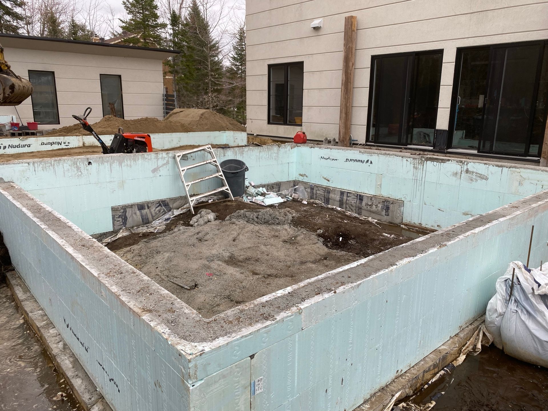 Chantier : piscine inachevée avec des coffrages en béton isolés bleu clair, entourée d'un bâtiment et d'un hangar.