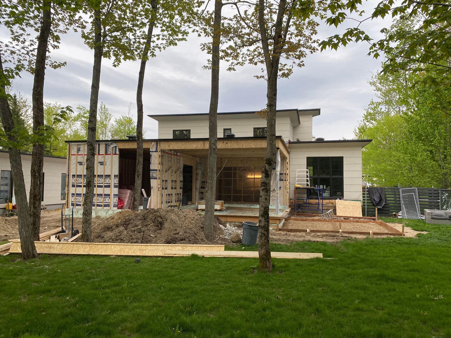 Maison en construction avec charpente apparente, entourée d'arbres et de pelouse verte.