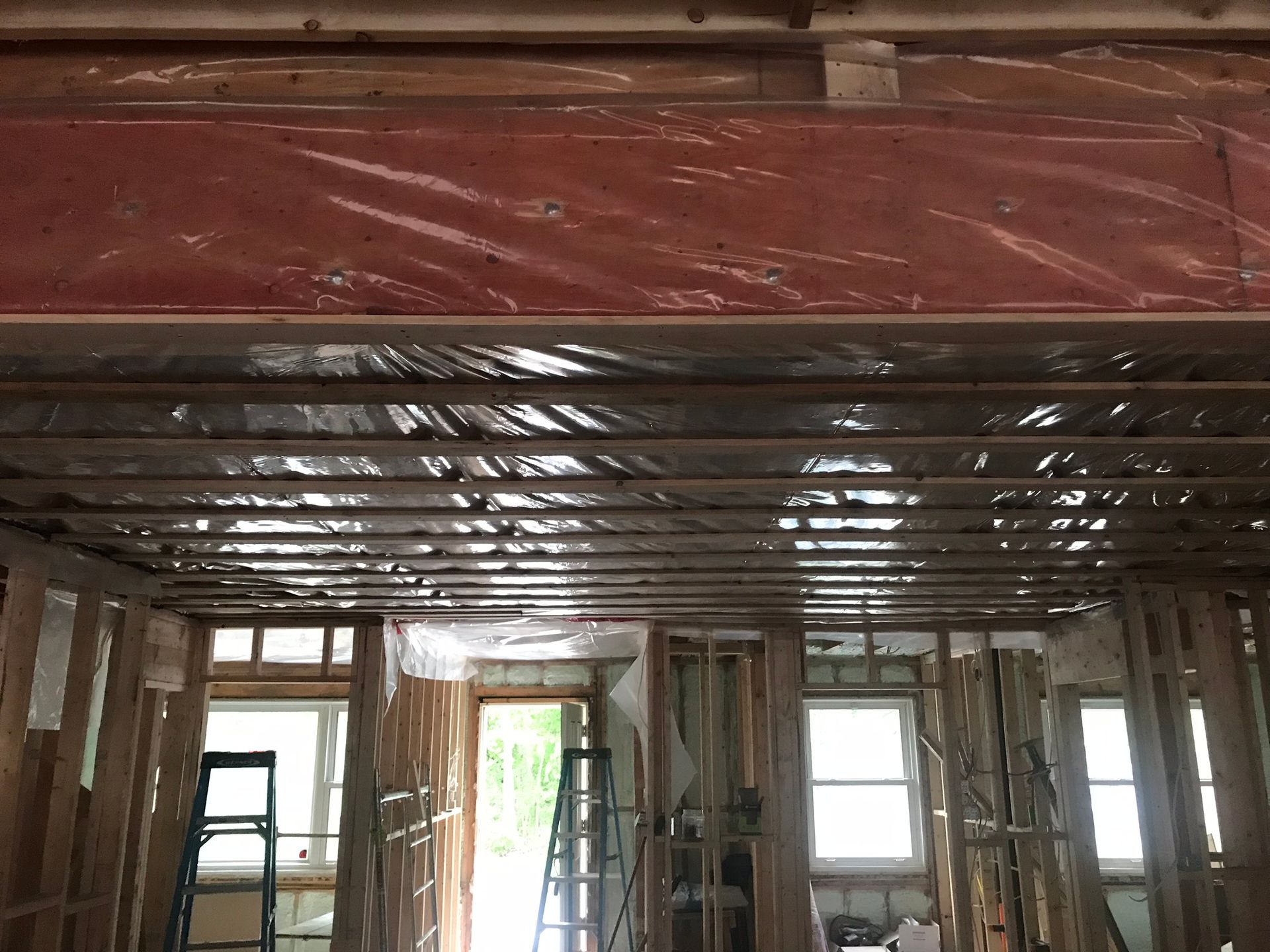 Intérieur d'une maison en construction : murs à ossature bois, bâches en plastique transparent recouvrant les plafonds et les poutres, lumière du soleil entrant par les fenêtres.