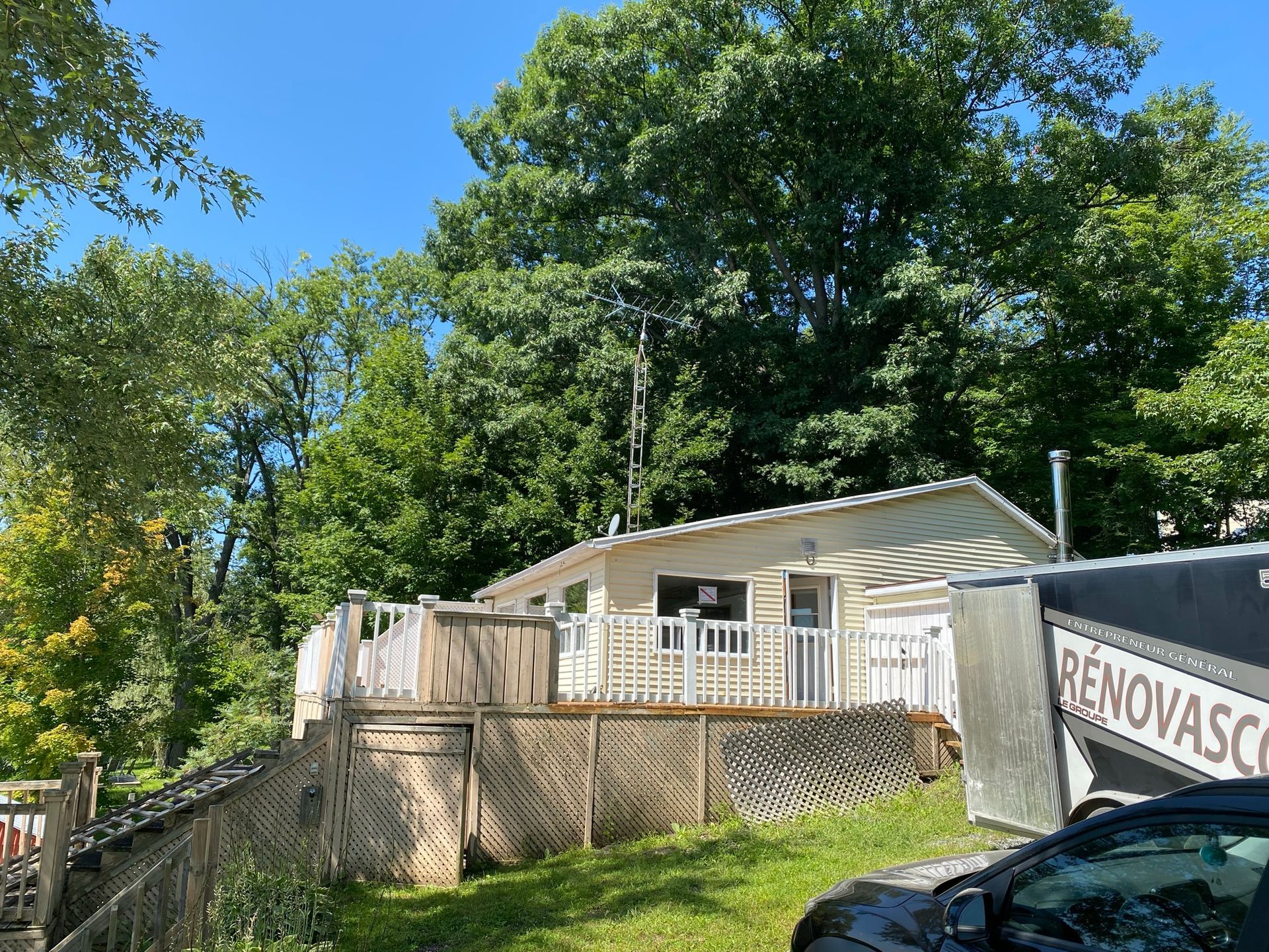 Petite maison blanche avec terrasse, entourée d'arbres par une journée ensoleillée.