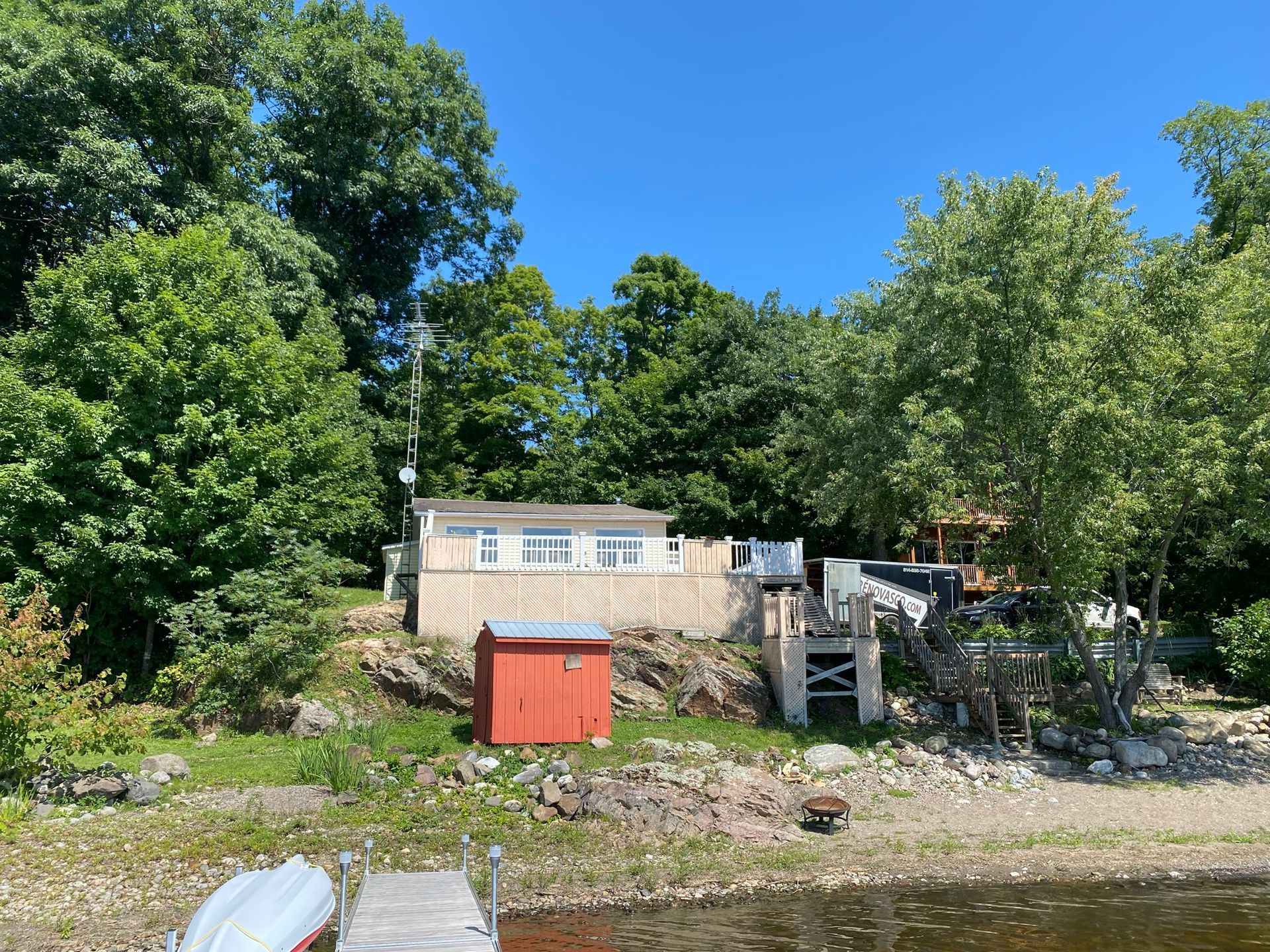 Chalet au bord d'un lac avec un petit abri et un escalier en bois, entouré d'arbres verdoyants sous un ciel bleu.
