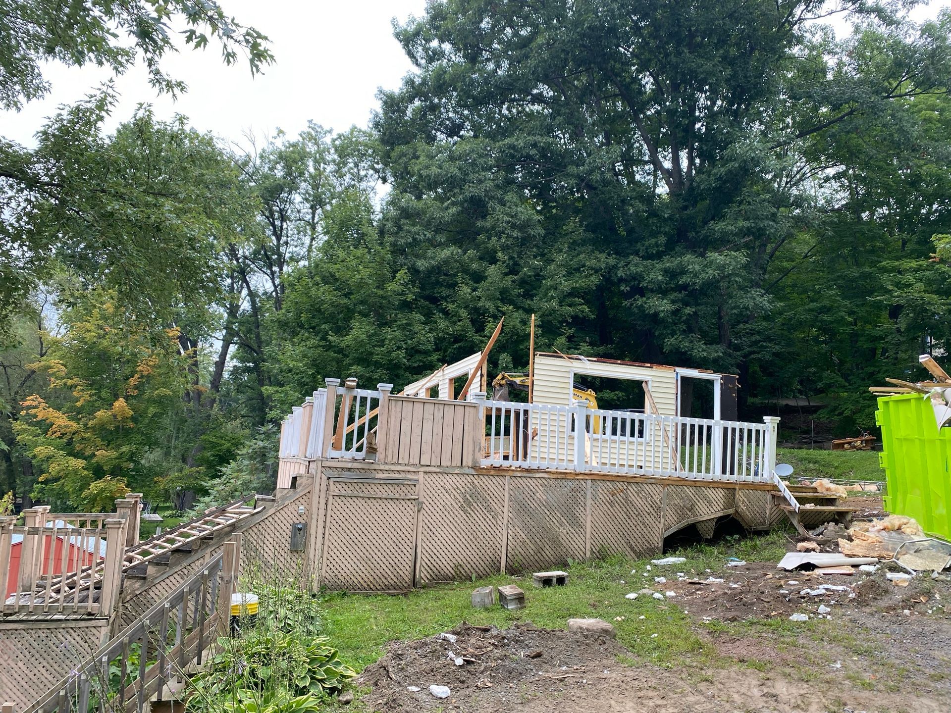 Une maison blanche endommagée, dotée d'une terrasse, se dresse dans une cour entourée d'arbres ; des débris sont éparpillés.