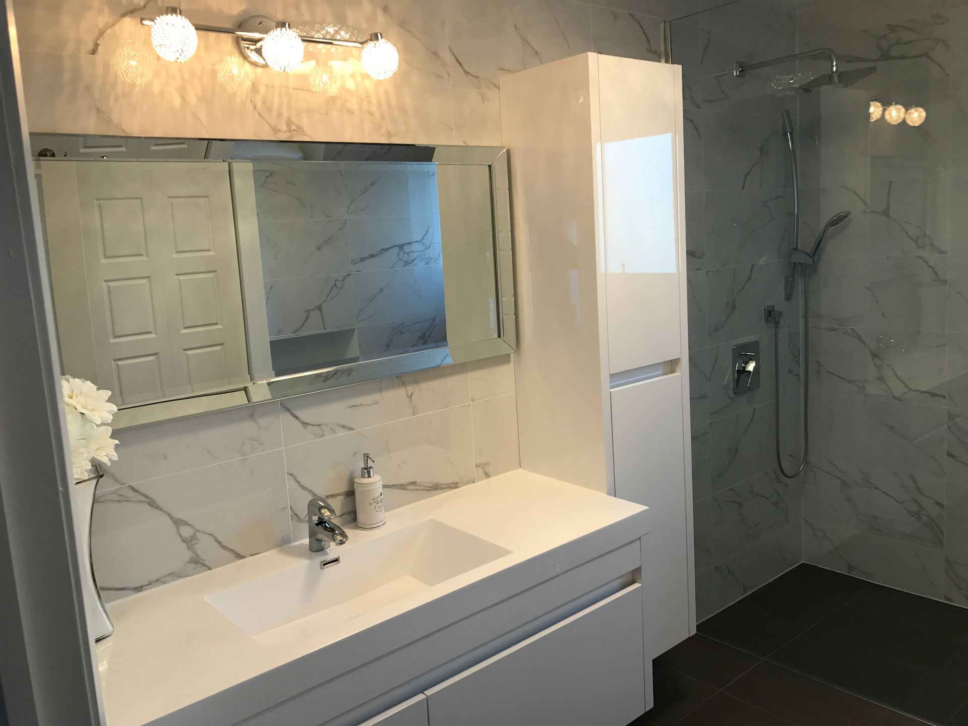 Salle de bains moderne avec meuble-lavabo blanc, miroir, douche et armoire de rangement haute sur un carrelage imitation marbre.