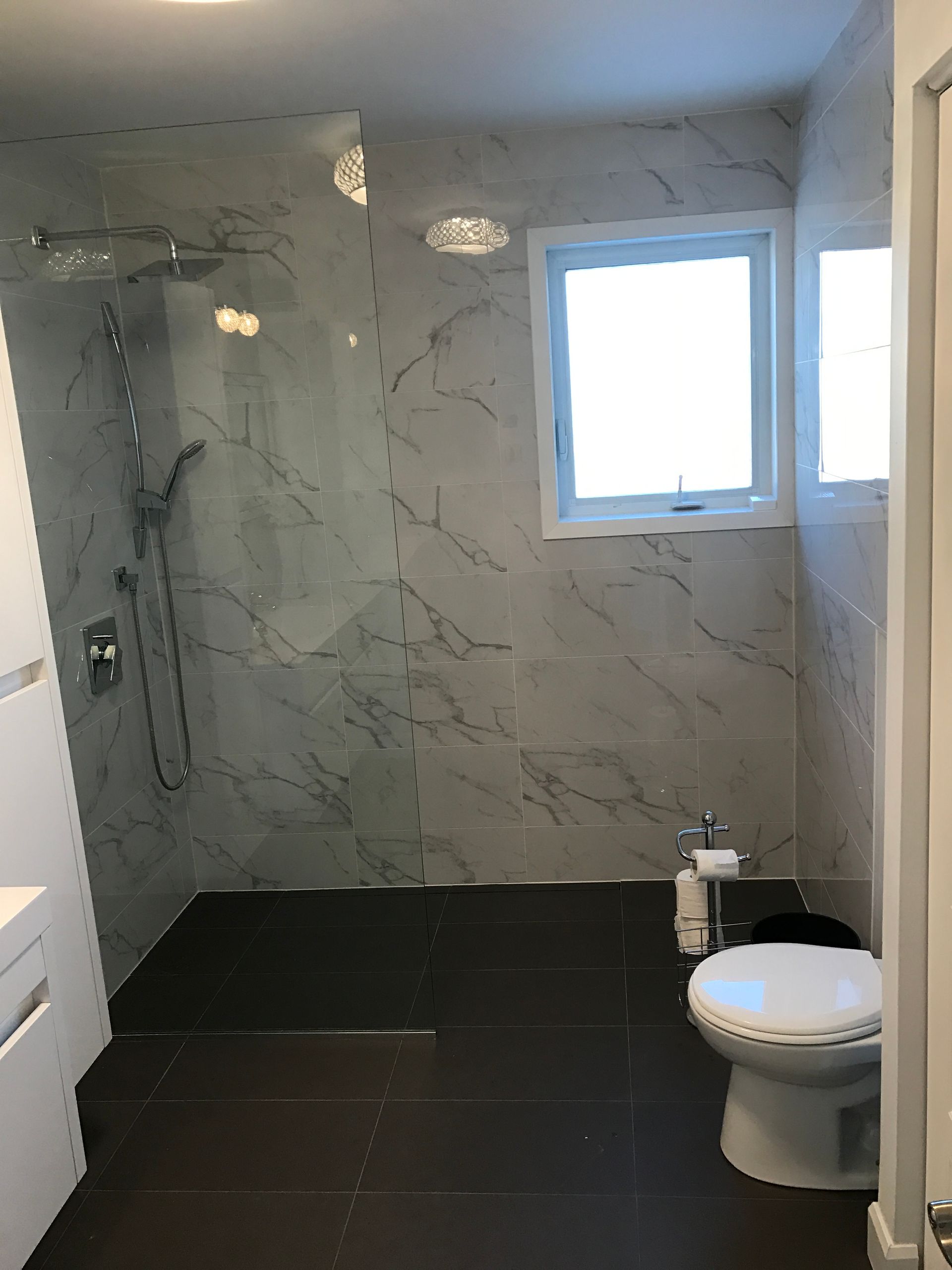 Salle de bains avec douche carrelée à motif marbre et sol foncé ; toilettes et fenêtre présentes.