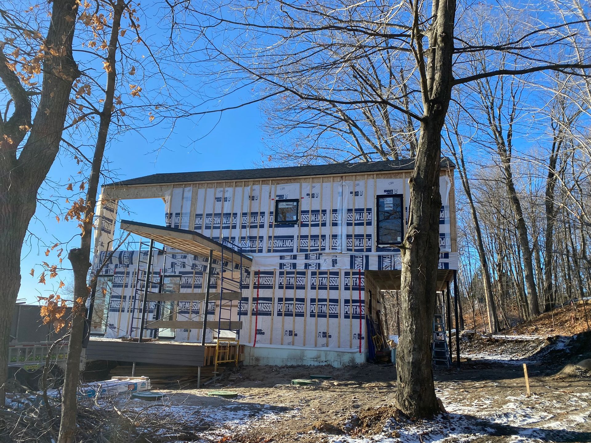 Construction d'une maison en hiver : ossature bois, bâche bleue, neige au sol, arbres et ciel dégagé.