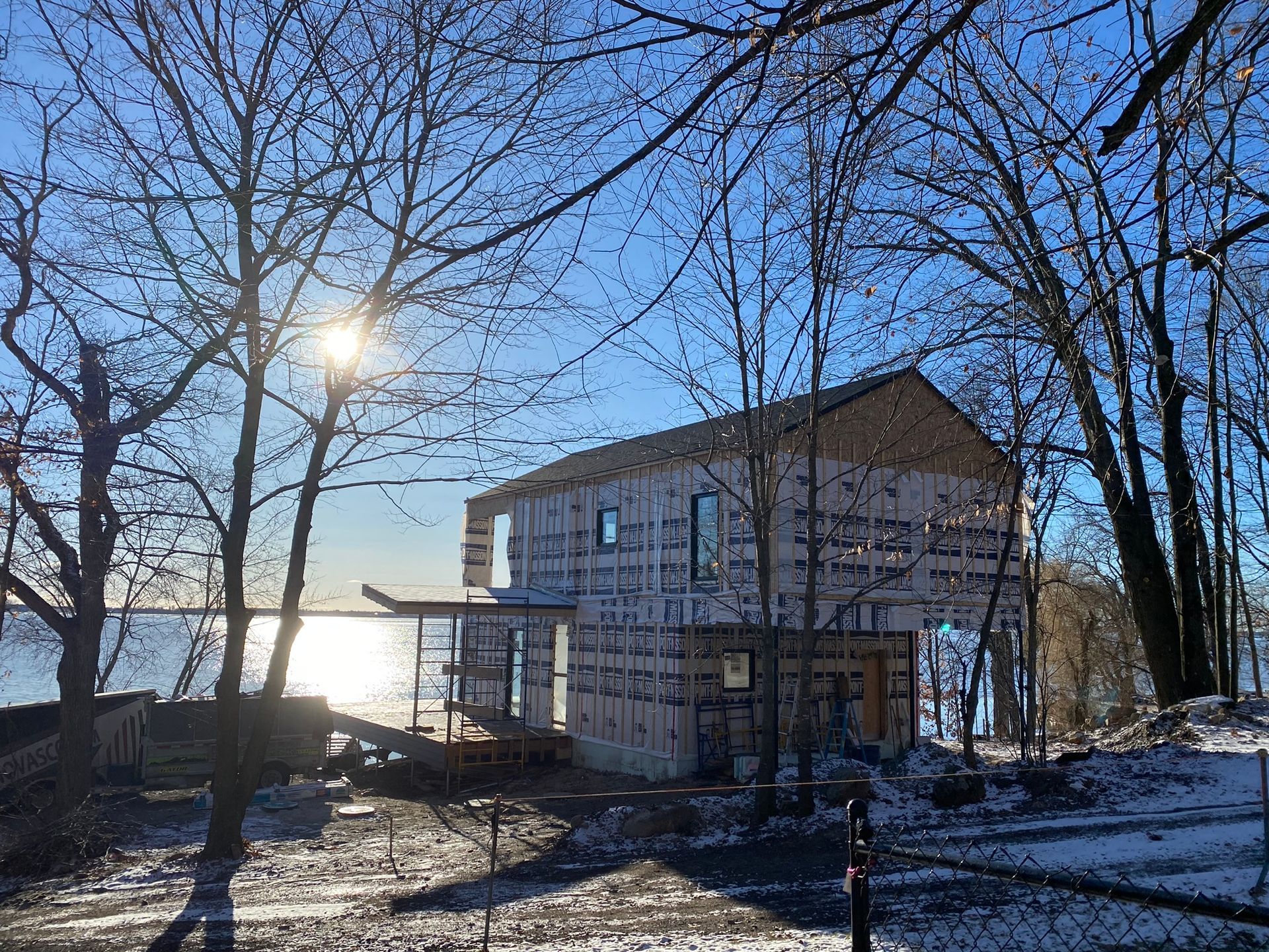 Maison à deux étages en construction sur une colline enneigée surplombant l'eau, arbres au premier plan, soleil éclatant.