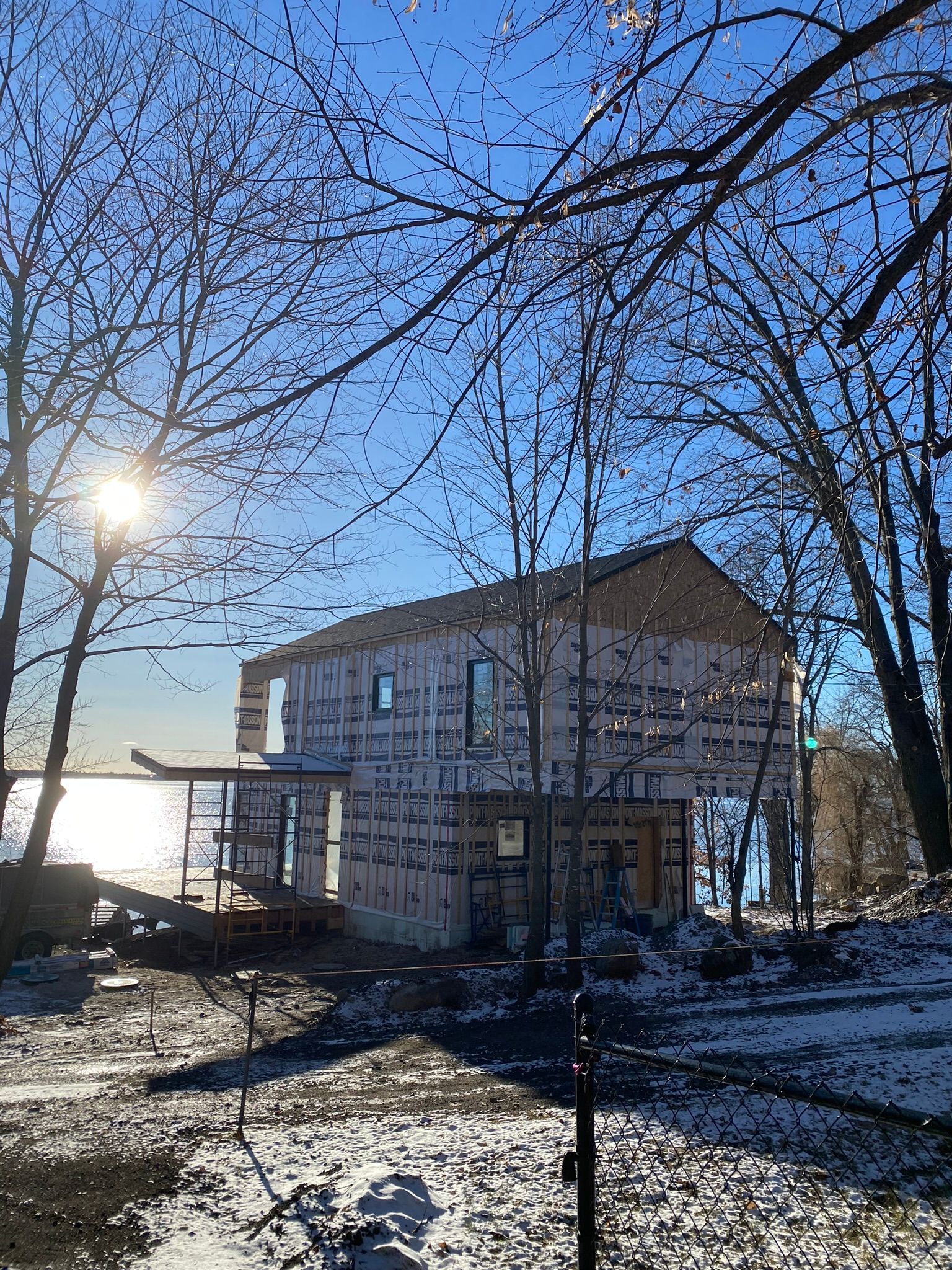 Bâtiment en construction par une journée enneigée, encadré par des arbres dénudés, avec un soleil éclatant et une étendue d'eau au loin.