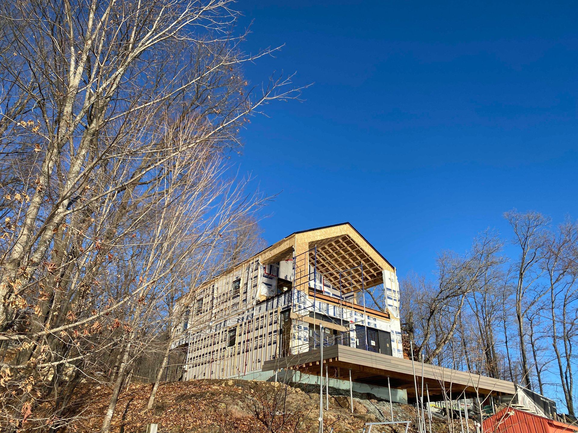 Maison en construction avec charpente et bardage apparents, se détachant sur un ciel bleu, avec des arbres au premier plan.