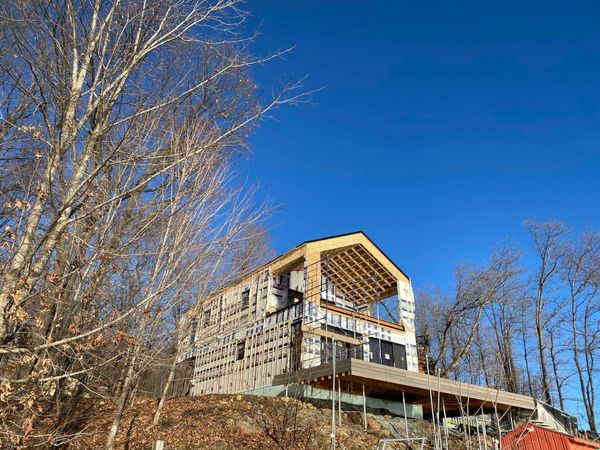 Maison en construction avec charpente et bardage apparents, se détachant sur un ciel bleu, avec des arbres au premier plan.