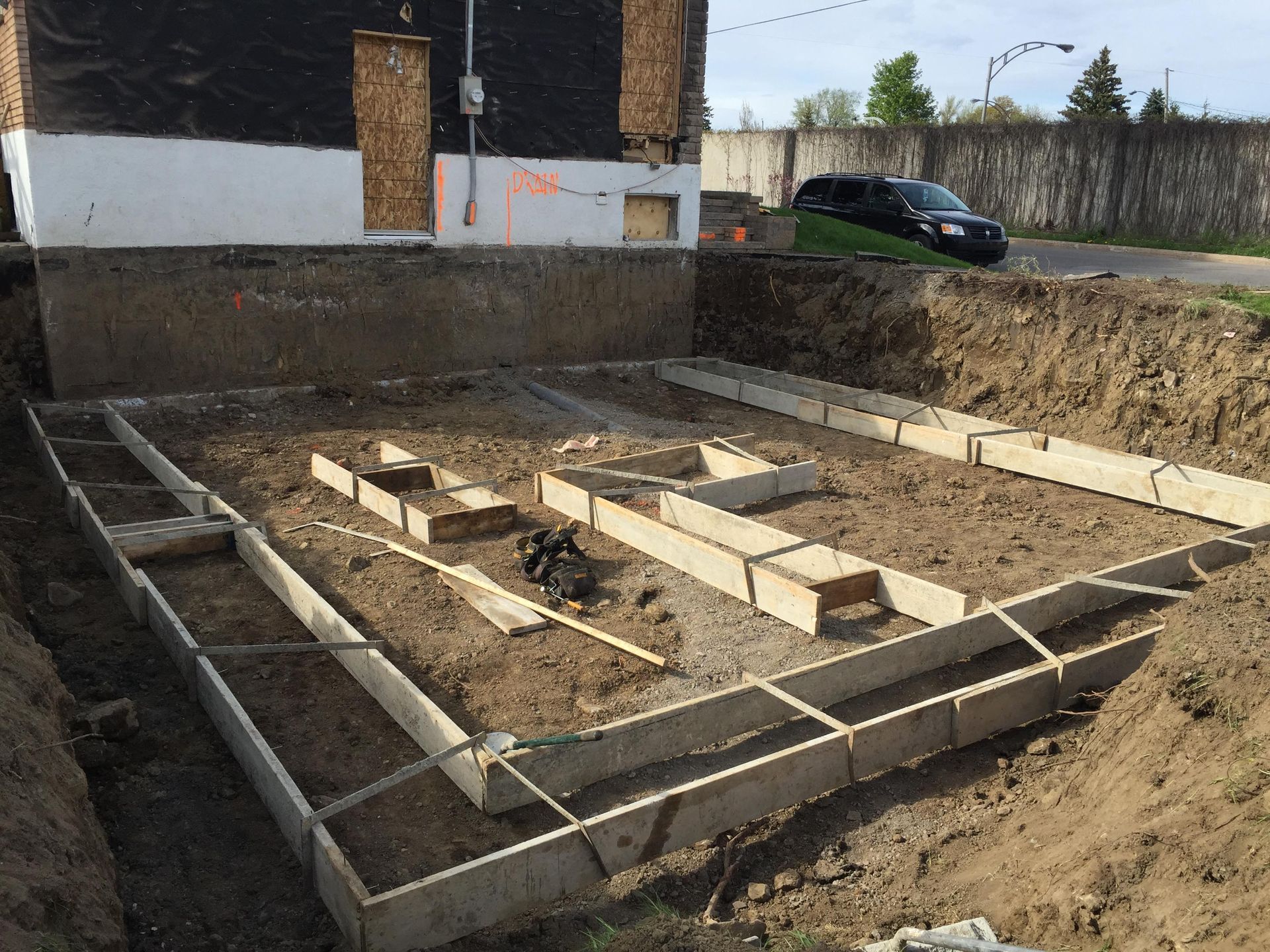 Chantier de construction : Coffrages de fondation dans une zone excavée à côté d'un bâtiment.