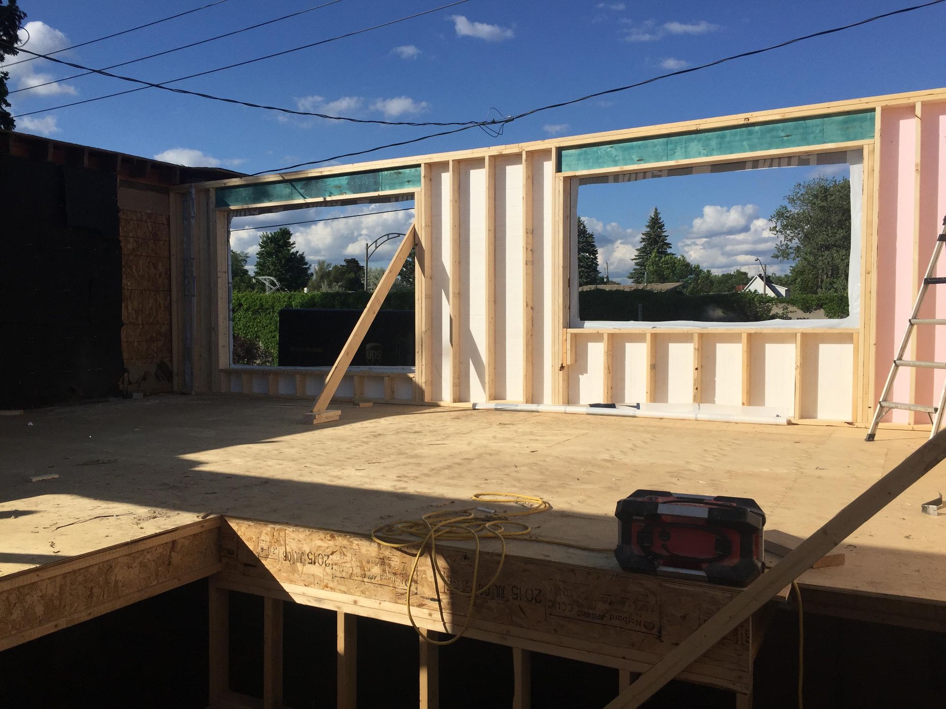 Chantier : Murs à ossature bois avec ouvertures pour fenêtres sous un ciel bleu ; sol inachevé et outils visibles.