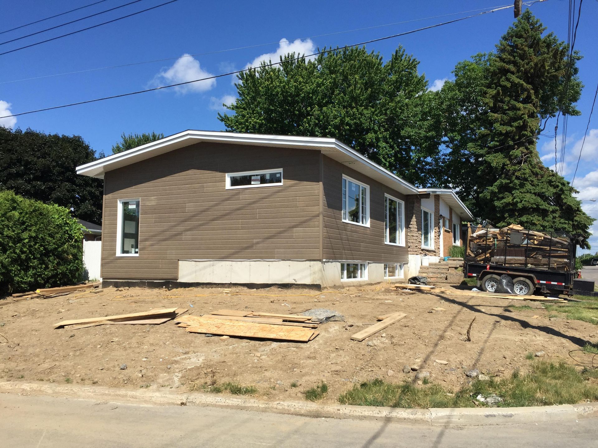 Maison en construction, façade brune et boiseries blanches. Une caravane chargée de bois est garée à côté, sur un terrain vague.