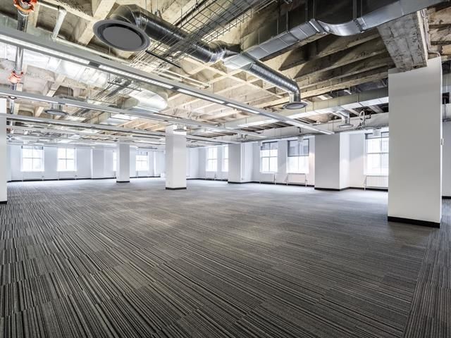 Espace de bureau vide avec moquette au sol, piliers blancs, fenêtres et luminaires apparents au plafond.
