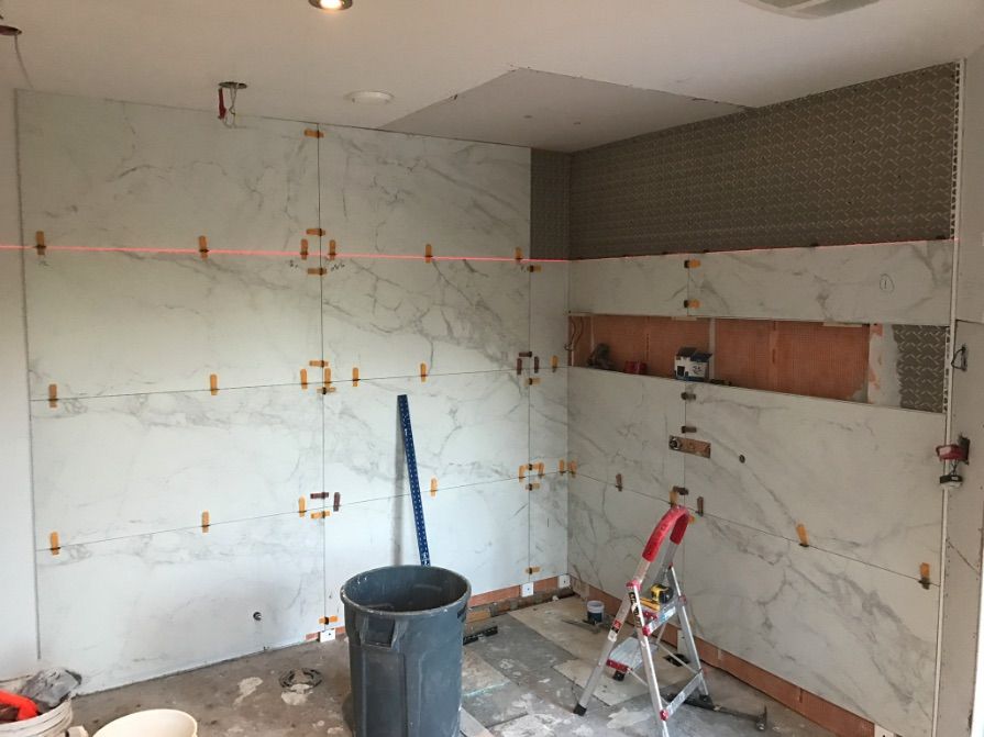 Salle de bains en travaux : pose de grands carreaux imitation marbre. Niveau laser utilisé ; poubelle, échelle et outils visibles.