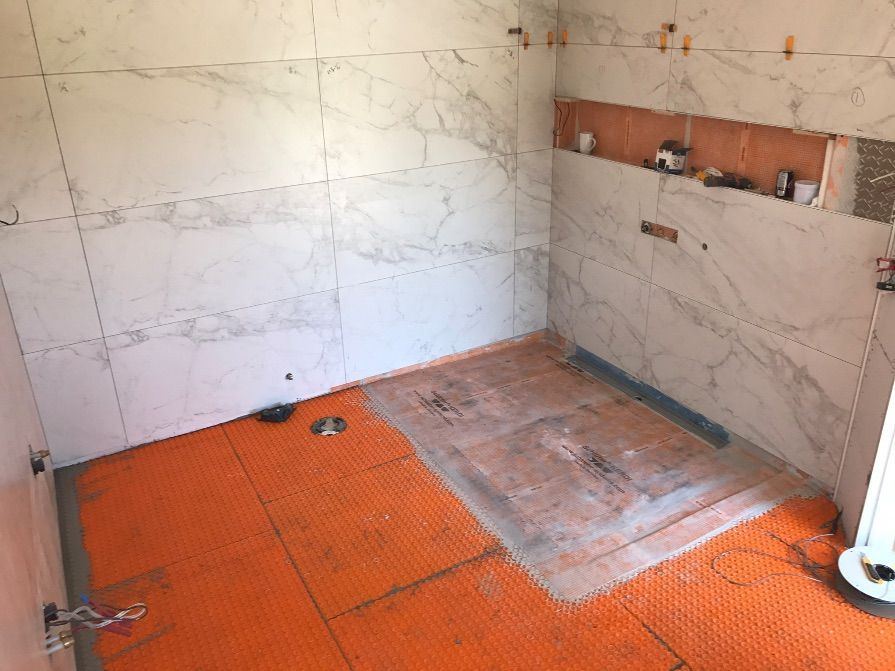 Salle de bains en cours de rénovation : murs et sol carrelés de grands carreaux blancs imitation marbre. Membrane d’étanchéité orange installée.