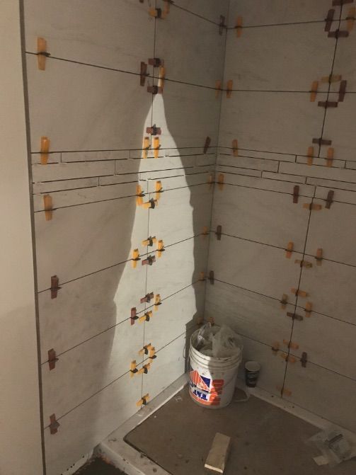 Coin de douche carrelé en cours de réalisation avec des cales de nivellement et un seau à l'intérieur. Tons blancs et beiges.
