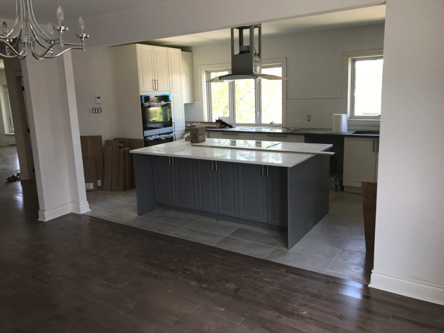 Cuisine en cours de rénovation ; îlot gris, comptoirs blancs, armoires claires et planchers en bois franc foncé.
