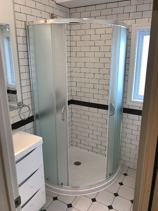 Salle de bains avec douche à porte vitrée, carrelage blanc imitation brique, bande décorative foncée et meuble-lavabo.
