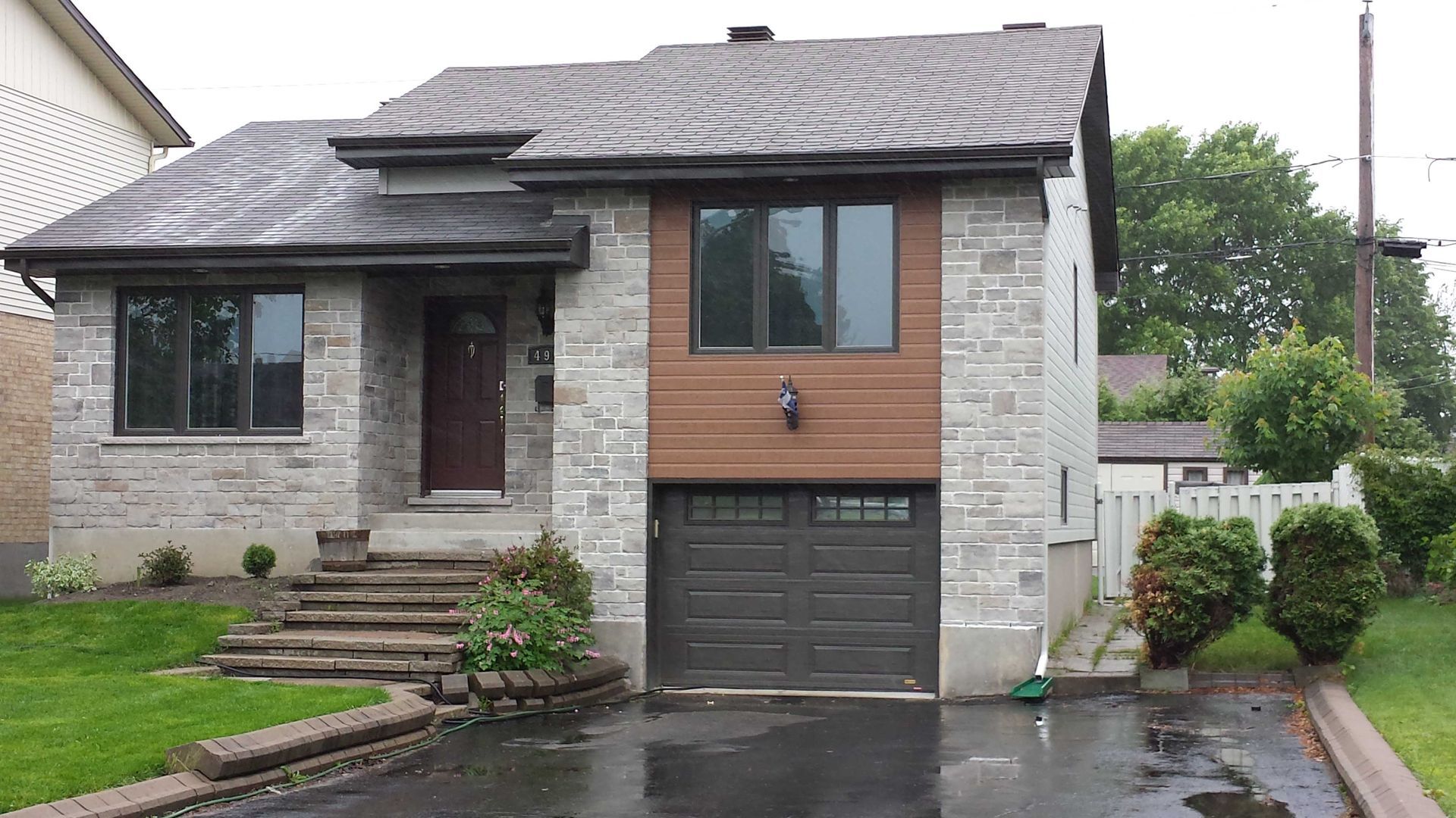 Une maison en briques de deux étages avec une porte de garage marron et une petite cour avant avec de l'asphalte mouillé.
