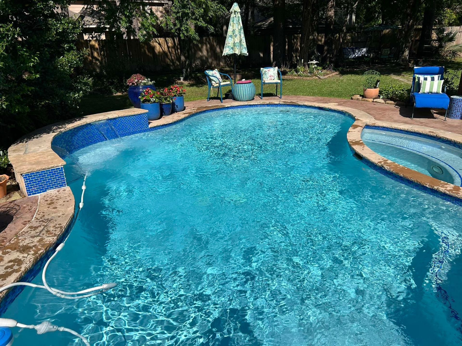 Swimming pool with blue tiles, lounge chairs, umbrella, and lush greenery in a backyard.
