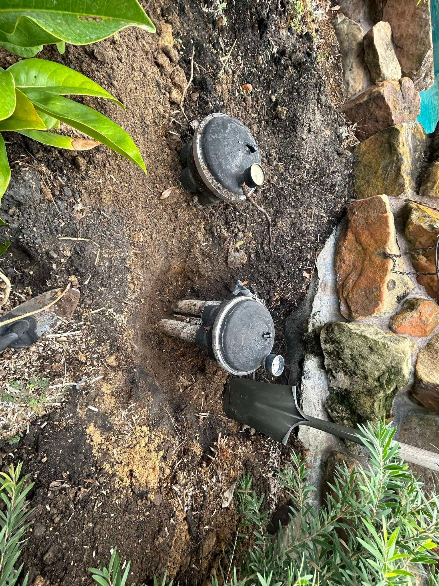 Two black sprinkler heads emerging from soil with a small shovel and surrounding greenery.