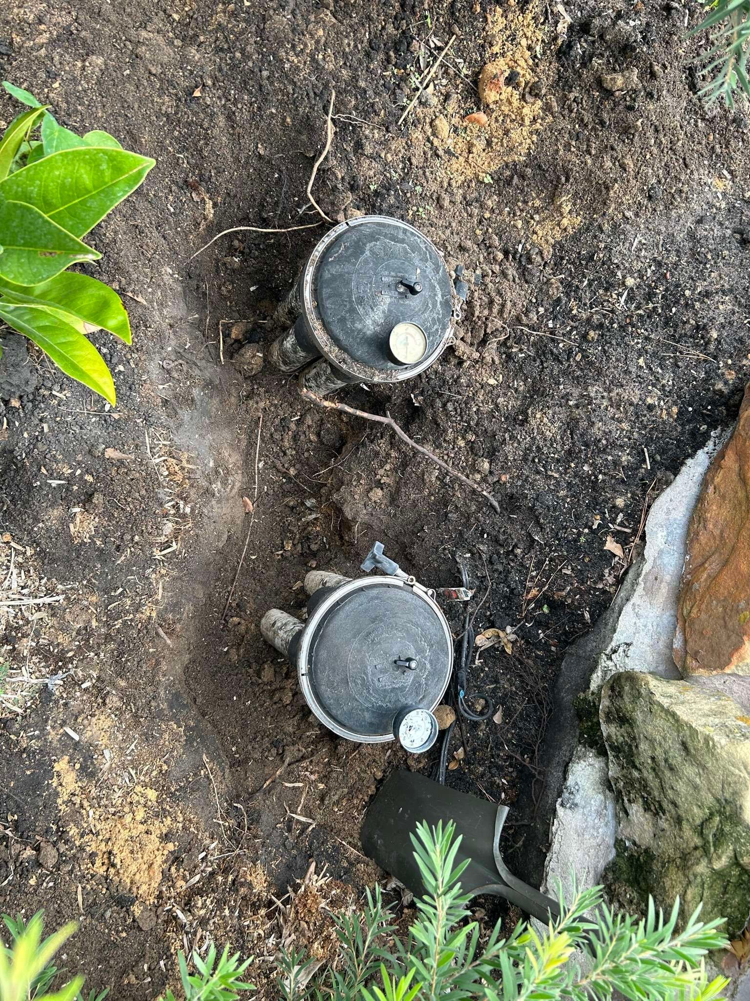 Two black sprinkler heads on brown soil with green plants nearby.