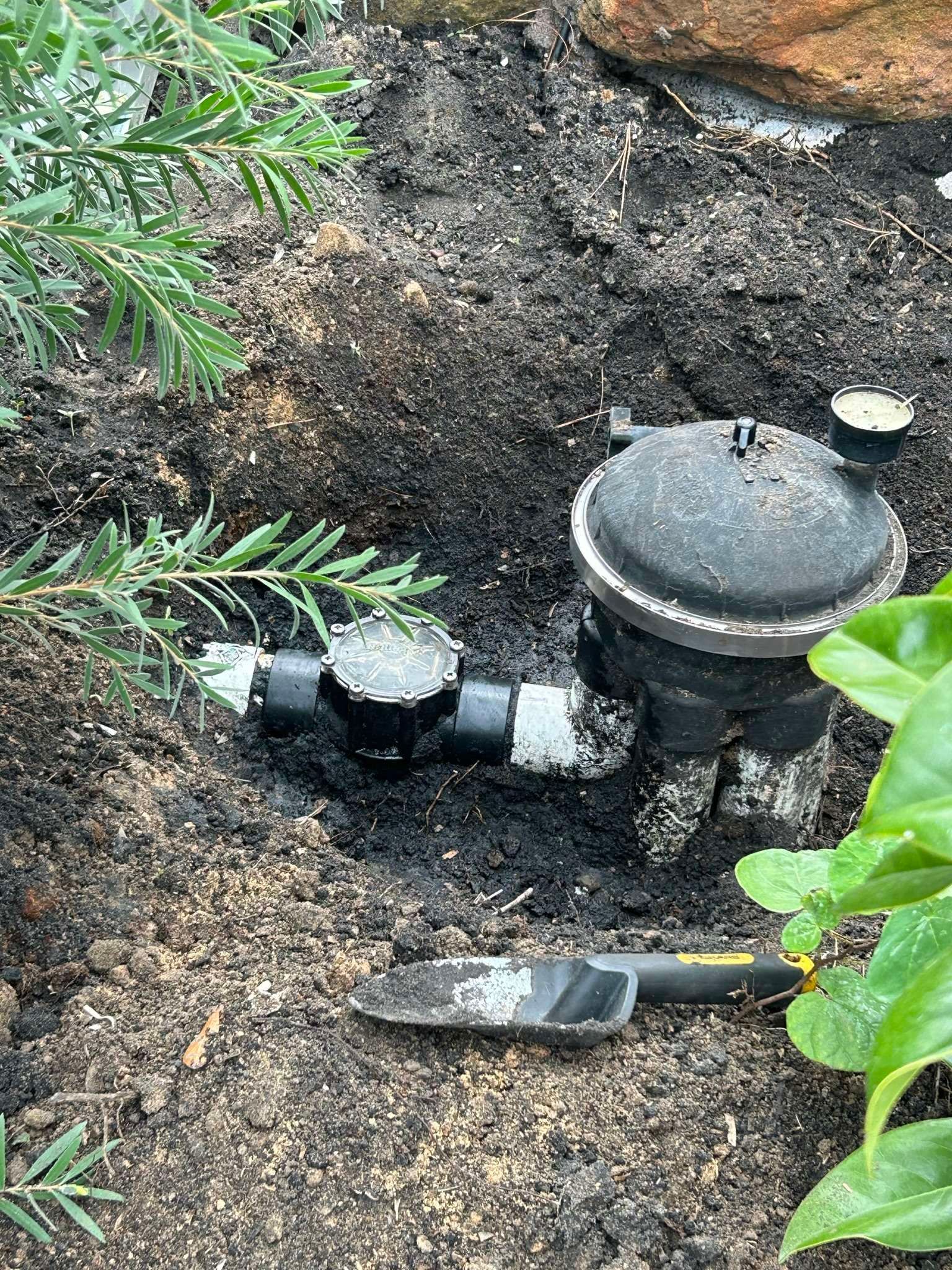 Black irrigation system components in dirt with a small shovel and green plants.