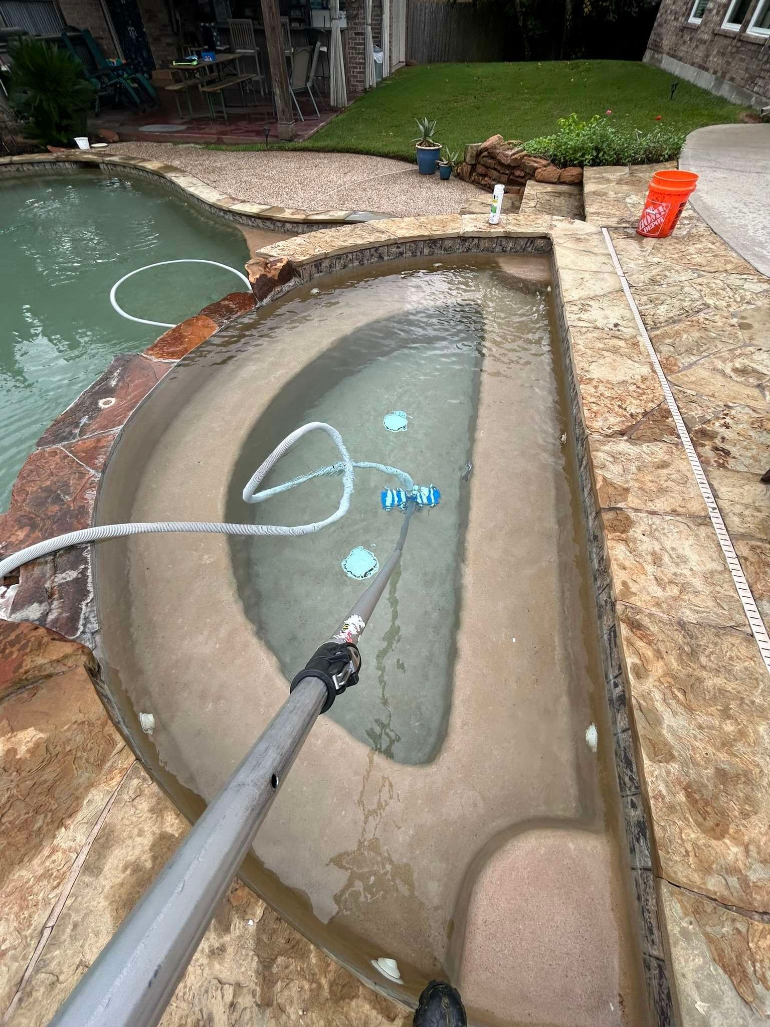 A person cleans a pool spa with a long-handled vacuum; green and brown stone surrounds.