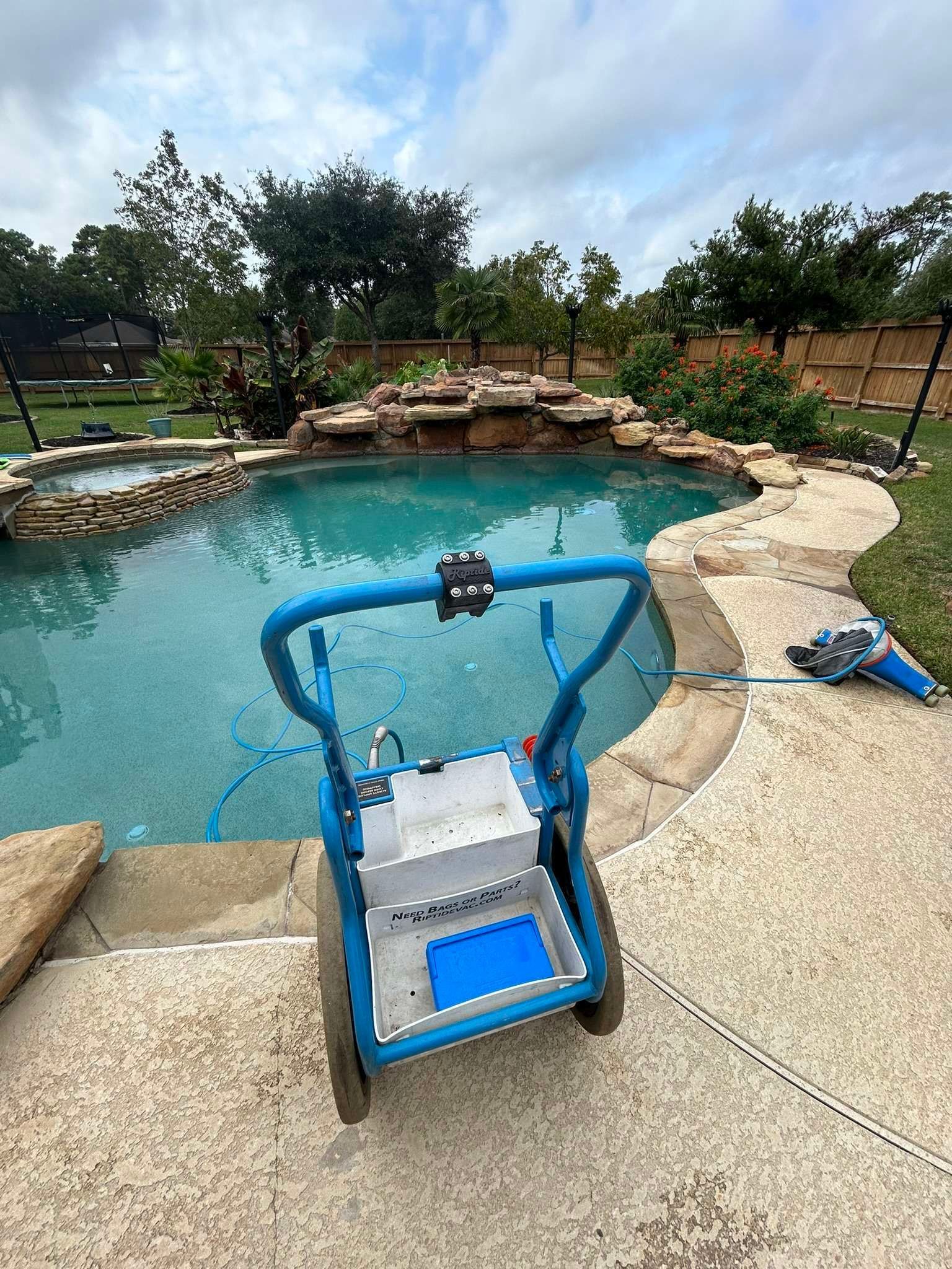 Blue pool cleaner on a pool deck next to a pool with a waterfall feature.
