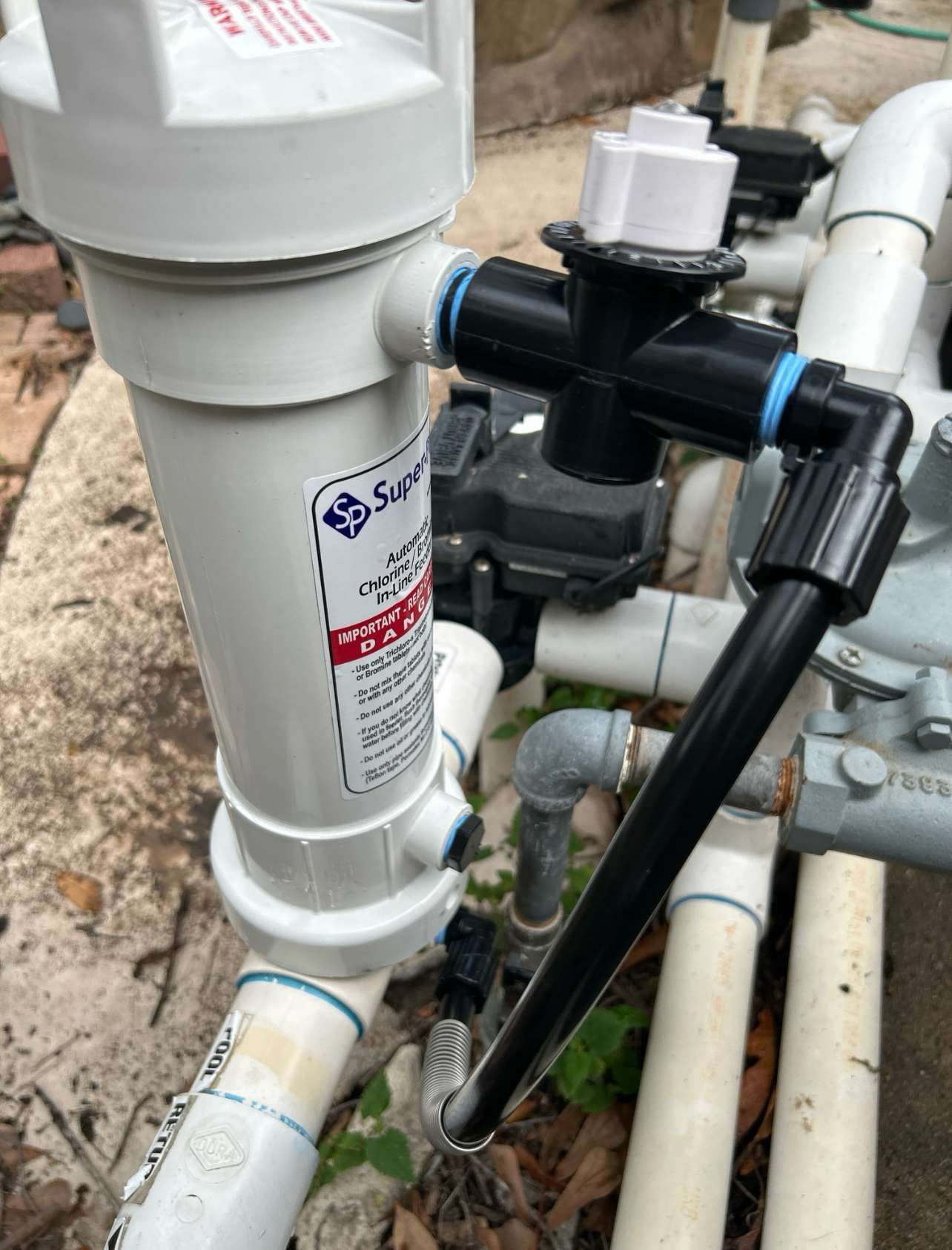 Pool filter system with white cylinder filter, black pipes, and a white valve, outdoors.