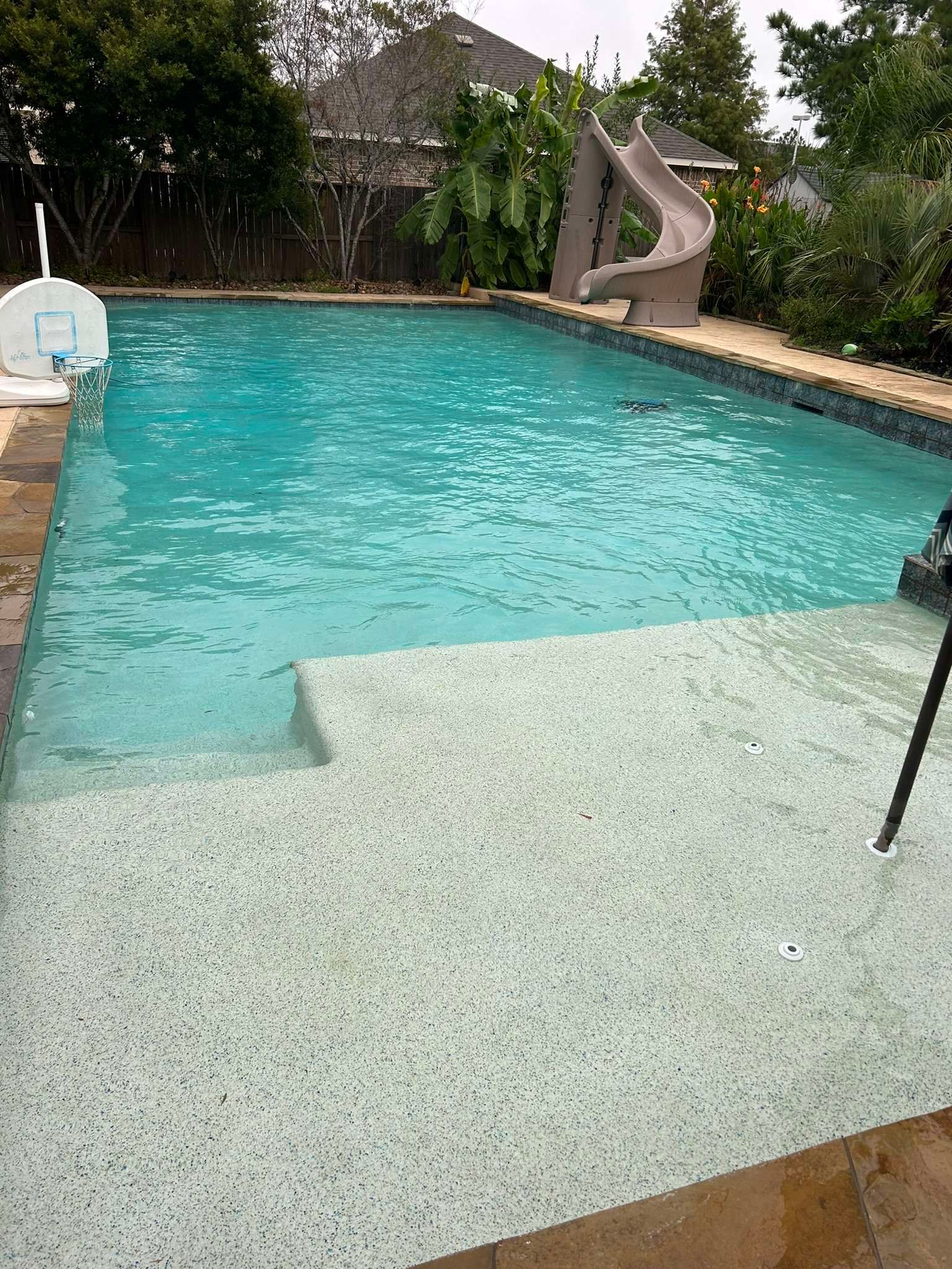 Pool with steps, slide, and basketball hoop in a backyard. Turquoise water reflects the sky.