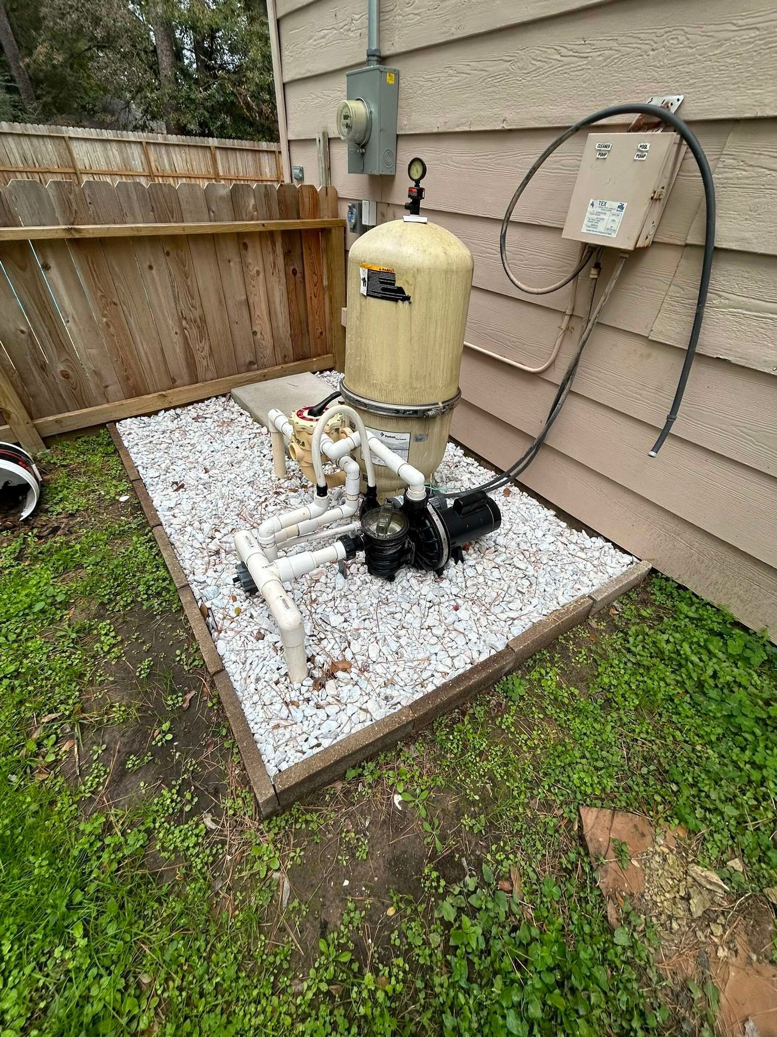 Pool filter system on a bed of white stones, next to a wooden fence and siding.