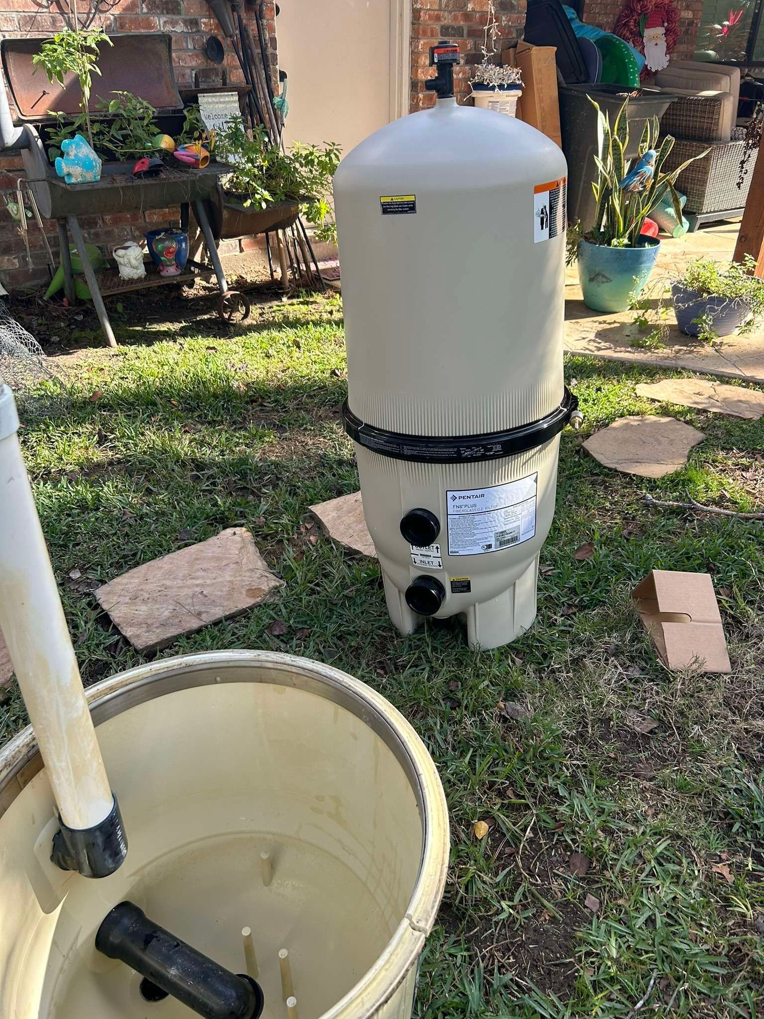 Pool filter with attached pipes sitting on grass near a bucket outdoors.