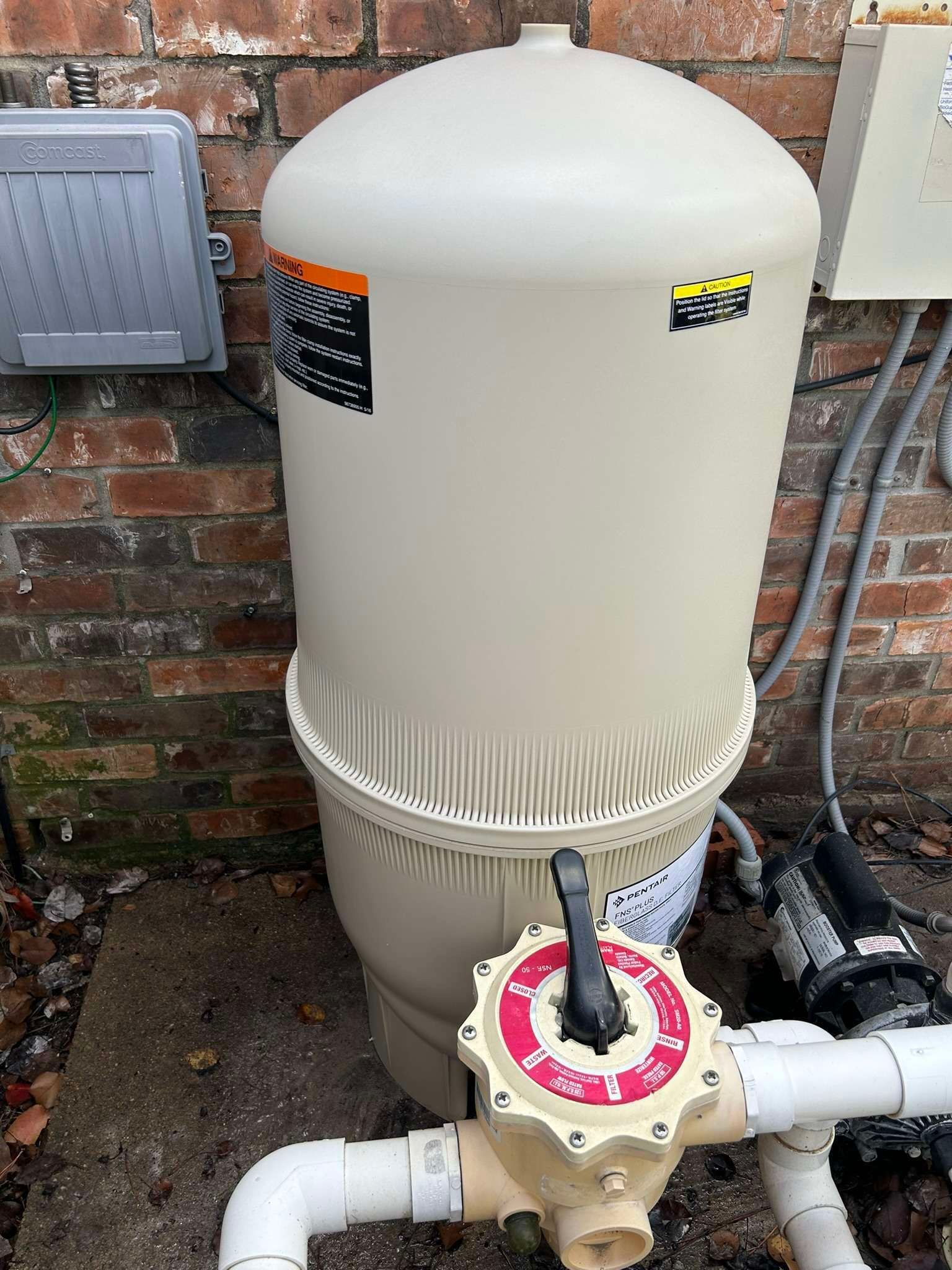 Tan pool filter with valve and pump against a brick wall.