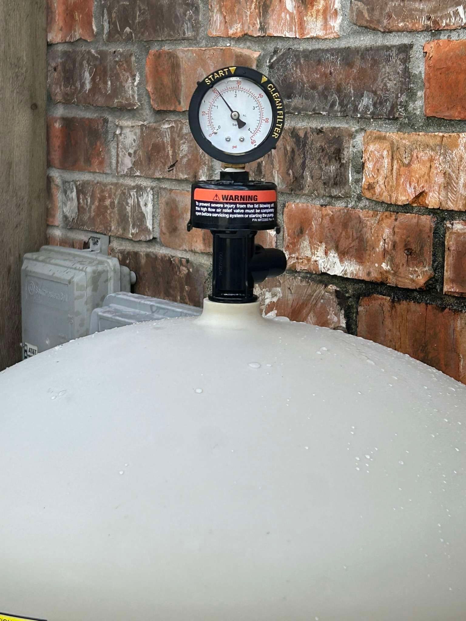 Pressure gauge on top of a white pool filter against a brick wall. The gauge reads pressure.