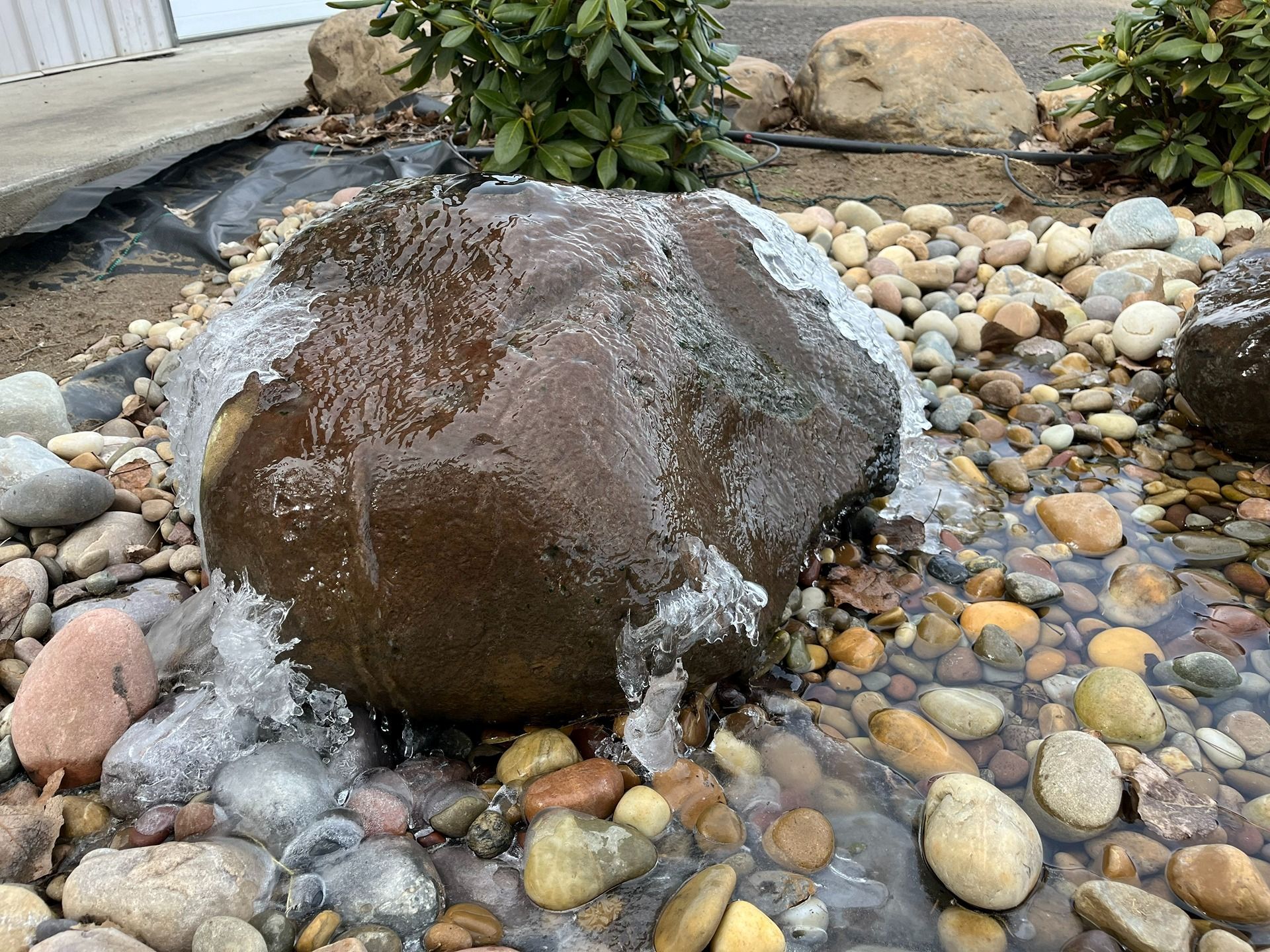 A large, dark rock fountain covered in thin, flowing water, surrounded by smooth decorative river stones.