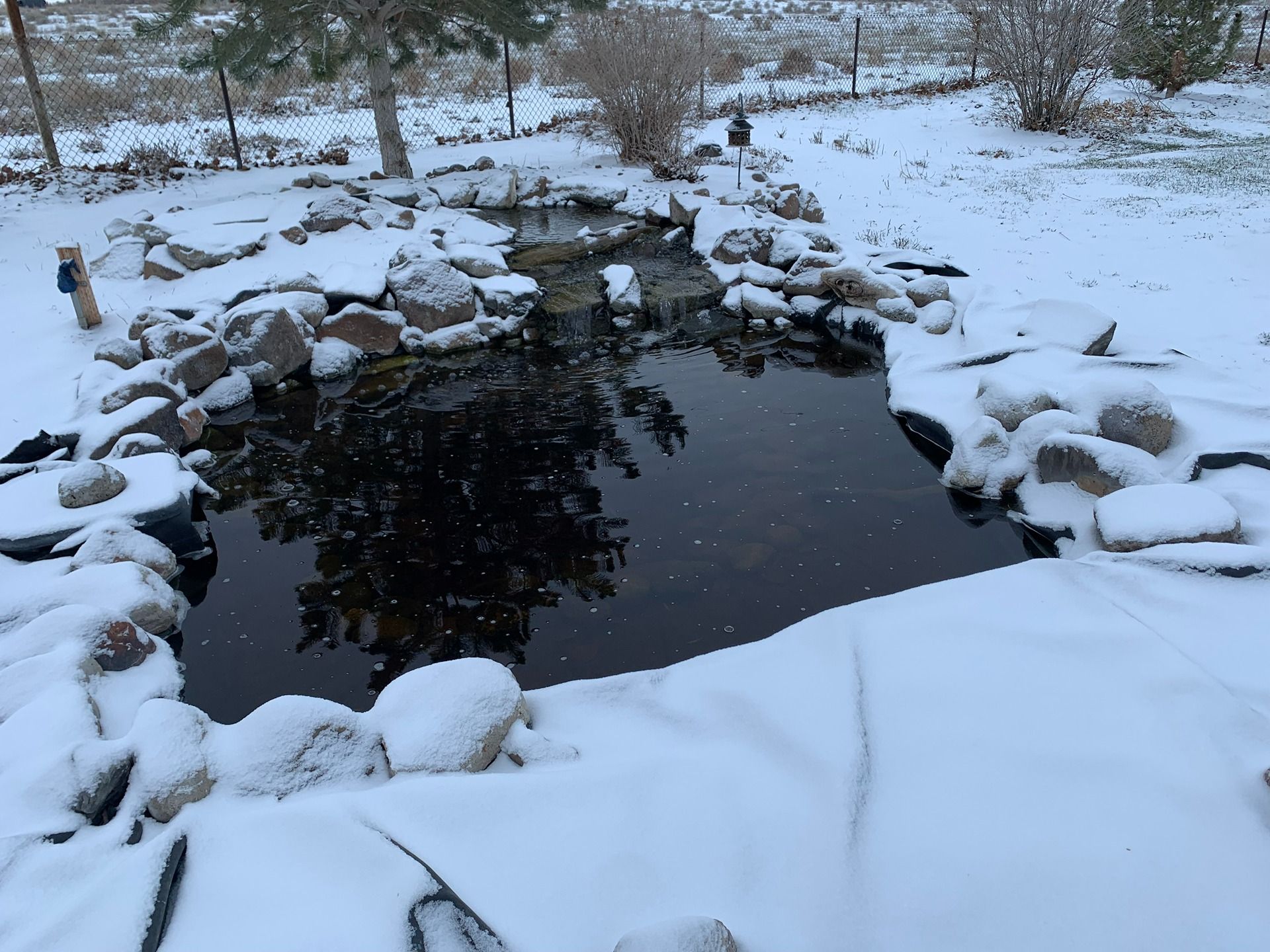 A snow-covered rock garden pond with a small waterfall feature in a winter landscape.