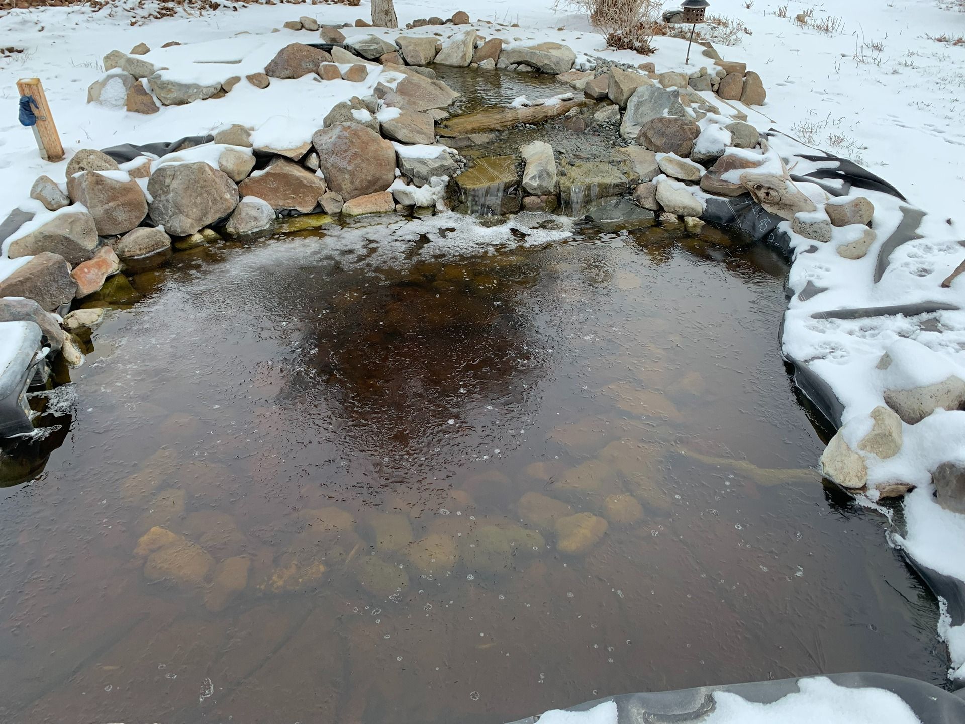 A stone-lined garden pond with a small waterfall, partially covered in snow and ice during winter.